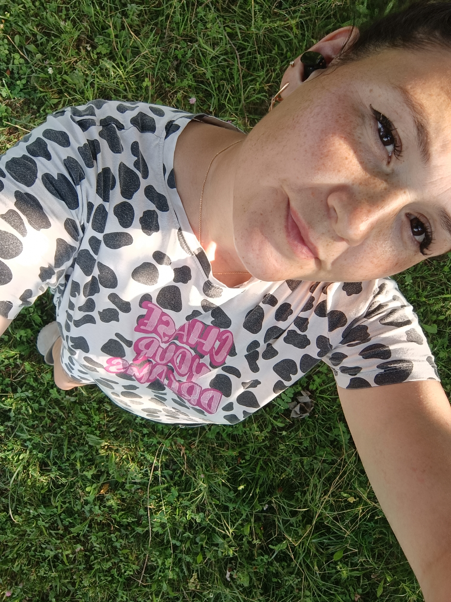 An overhead shot captures a woman lying on a grassy area, likely a park or backyard, with her face tilted towards the camera. Her face is illuminated by sunlight, casting a shadow across her forehead and cheek. She has fair skin with noticeable freckles. Her eyes are dark and she appears to be wearing mascara and eyeliner. Her lips are slightly parted, and she has a subtle smile. She's wearing a white t-shirt with a black cow-print pattern and pink lettering on the front. A delicate gold necklace is visible around her neck. The background is dominated by green grass with some clover and scattered small leaves. The image does not contain any information that would allow for its location to be identified as Karsha, Russia.
