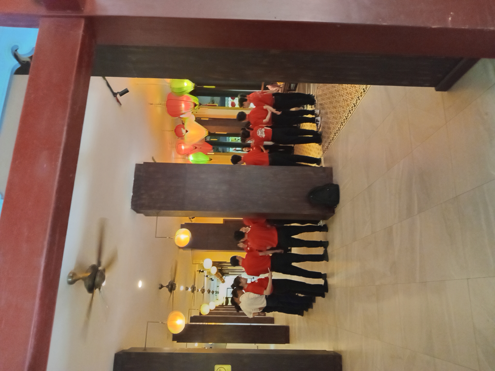 In a brightly lit indoor hallway with a high ceiling, a group of approximately ten individuals, mostly dressed in matching red shirts and dark pants, are gathered. Several ceiling fans are visible, along with decorative lanterns in vibrant colors like red, green, and yellow hanging from the ceiling. The floor is tiled with light-colored square tiles, bordered by a decorative patterned strip. The scene appears to be a professional or service setting, possibly a hotel or restaurant in Đà Nẵng, Vietnam, given the ambiance and cultural elements like the lanterns.