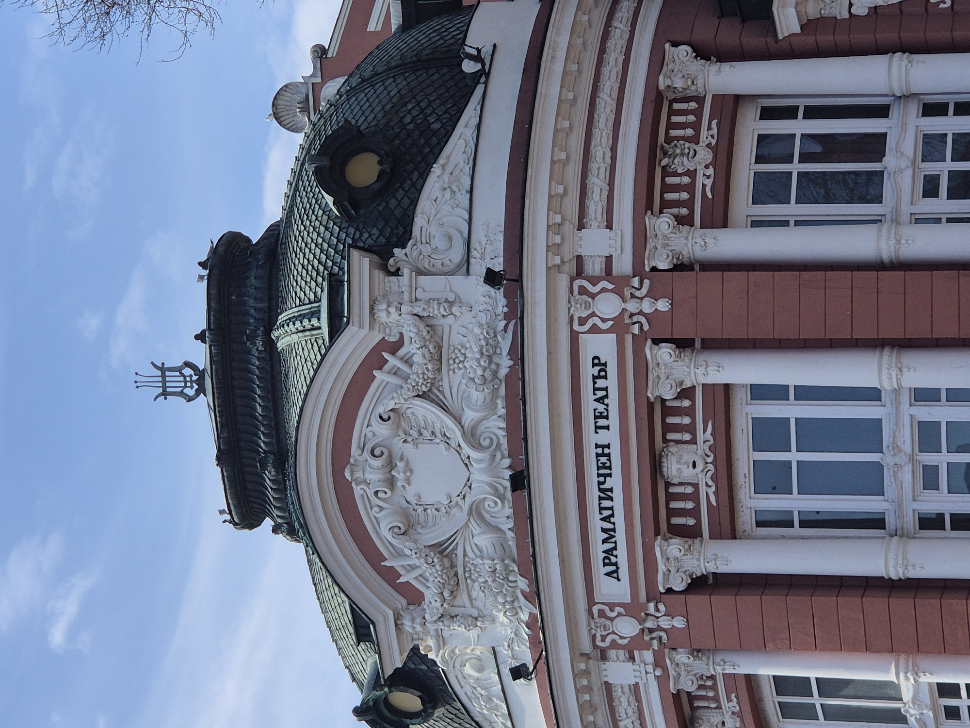 The image depicts a portion of the Ivan Vazov National Theatre in Sofia, Bulgaria. The architecture is a mix of Neo-Baroque and Neo-Classical styles, featuring a prominent dome with green tiles and ornate decorations. Below the dome is a section of the building with decorative white carvings and red-painted walls. Windows with white frames and classical columns are visible, along with the inscription "ДРАМАТИЧЕН ТЕАТЪР," which translates to "Drama Theatre" in Bulgarian. The sky is blue and clear. Bare branches of trees are visible in the upper left corner of the image.