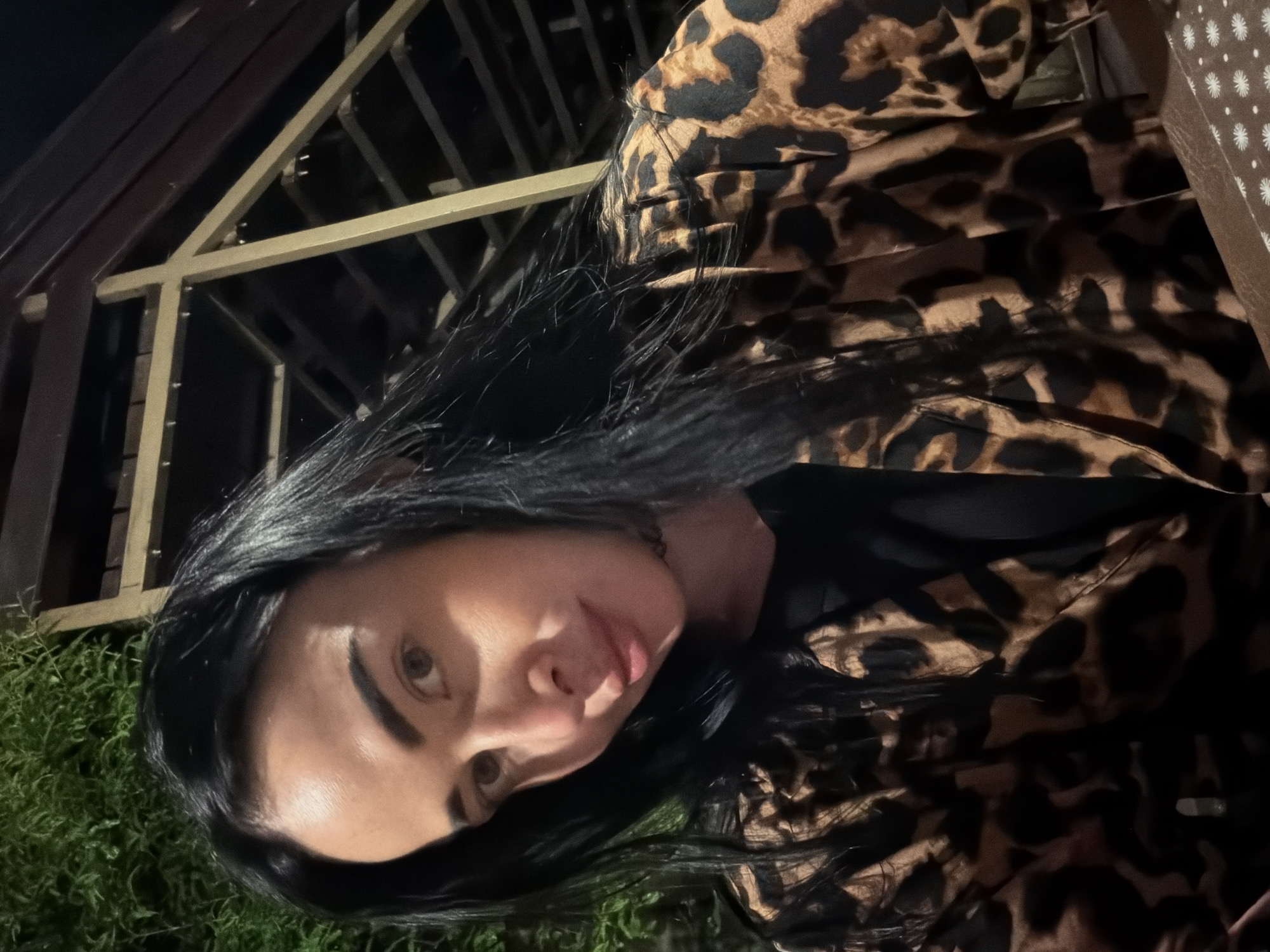 A medium shot selfie captures a woman with long, dark hair and light brown eyes. She is wearing a black shirt and a leopard-print jacket. The background features lush green foliage and a dark, structured roof with wooden and gold-colored beams. The lighting suggests it is evening or nighttime. Based on metadata, this image was taken in Sochi, Russia.