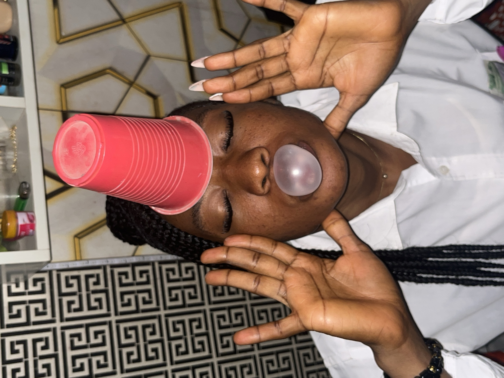 The media file shows a close-up, overhead shot of a young Black person lying down, with their head tilted back. The person is wearing a white collared shirt. Their eyes are closed, and they have a bubble of pink chewing gum inflated in front of their mouth. A red plastic cup is placed on their forehead, partially obscuring their face. Their hands are raised, palms facing upwards, with their fingers splayed. One hand is positioned above their forehead, near the cup, and the other is near their chest. The person has long, dark braided hair.
The background consists of a tiled floor with a black and white geometric pattern. Behind the person, a wall or surface with a gold-lined geometric design is visible. To the left, a shelf unit with various items, including what appear to be containers and a strand of beads, can be seen.
The lighting suggests an indoor setting, possibly a home or studio. There is no clear indication of the time of day or weather. The overall mood is playful and relaxed. No visible text is present in the media file. The context provided indicates the location is Jos, Nigeria.