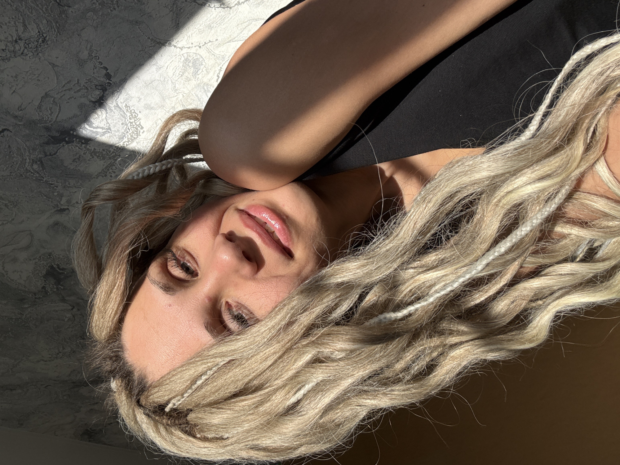 The image shows a woman lying on her side, likely indoors, with her head resting on her arm. She has light skin, blonde hair with white braid accents, and is wearing a dark top. Light from a window illuminates part of her face and the wall behind her, creating contrasting light and shadow patterns on the textured wall surface. The location, Slobodka (Kuznetsovskii S/O), Russia, is extraneous information as the image does not depict anything that confirms it.