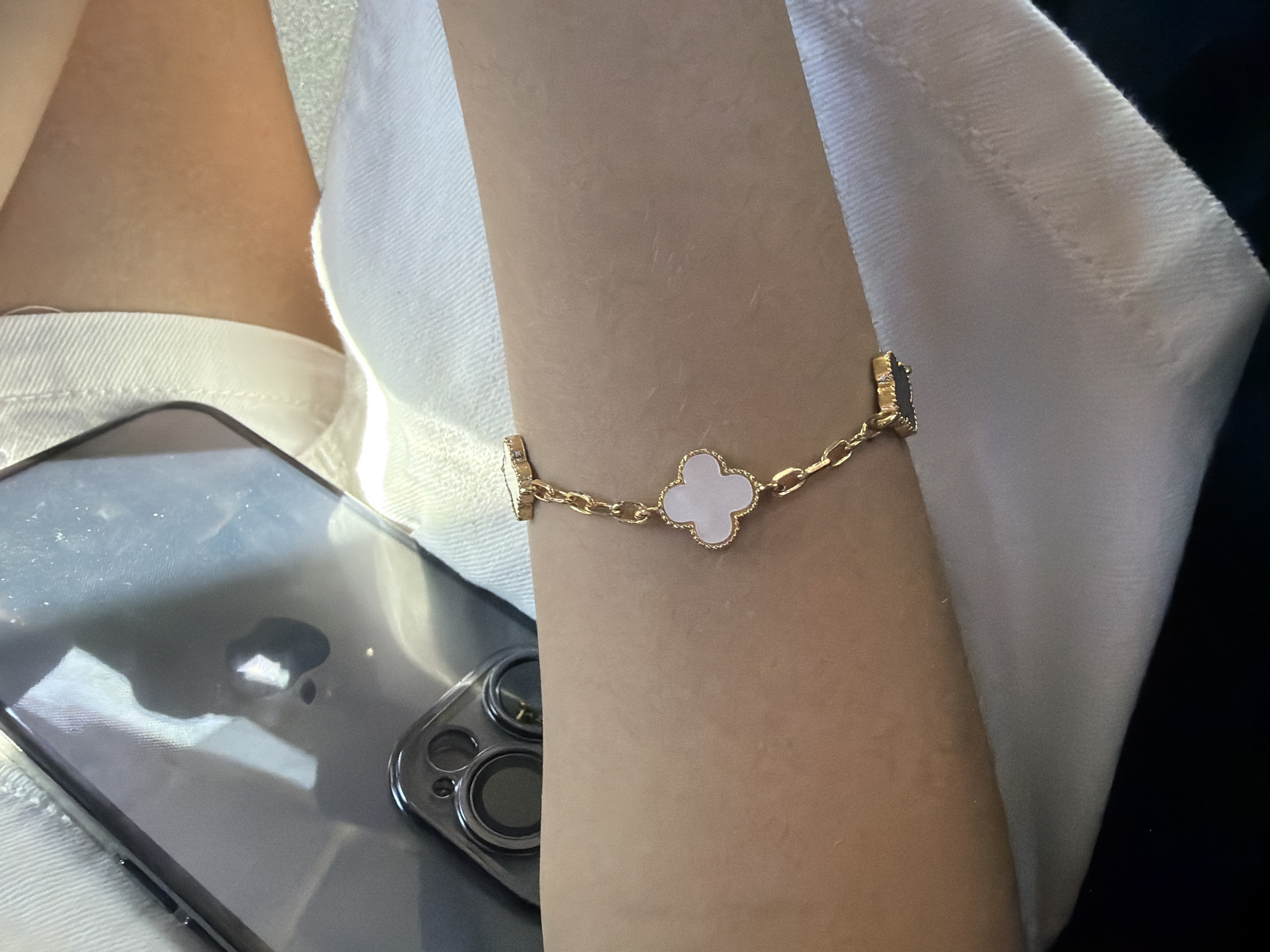 The image shows a close-up view of a person's arm wearing a gold bracelet with a clover-shaped charm made of white material. The arm is resting on a white textured surface, possibly clothing. In the upper left portion of the image, a dark smartphone with a visible camera bump is partially visible. The background is blurry and appears to be indoors. There is no identifiable information in the image that would allow for the determination of its location in Yerevan, Armenia.