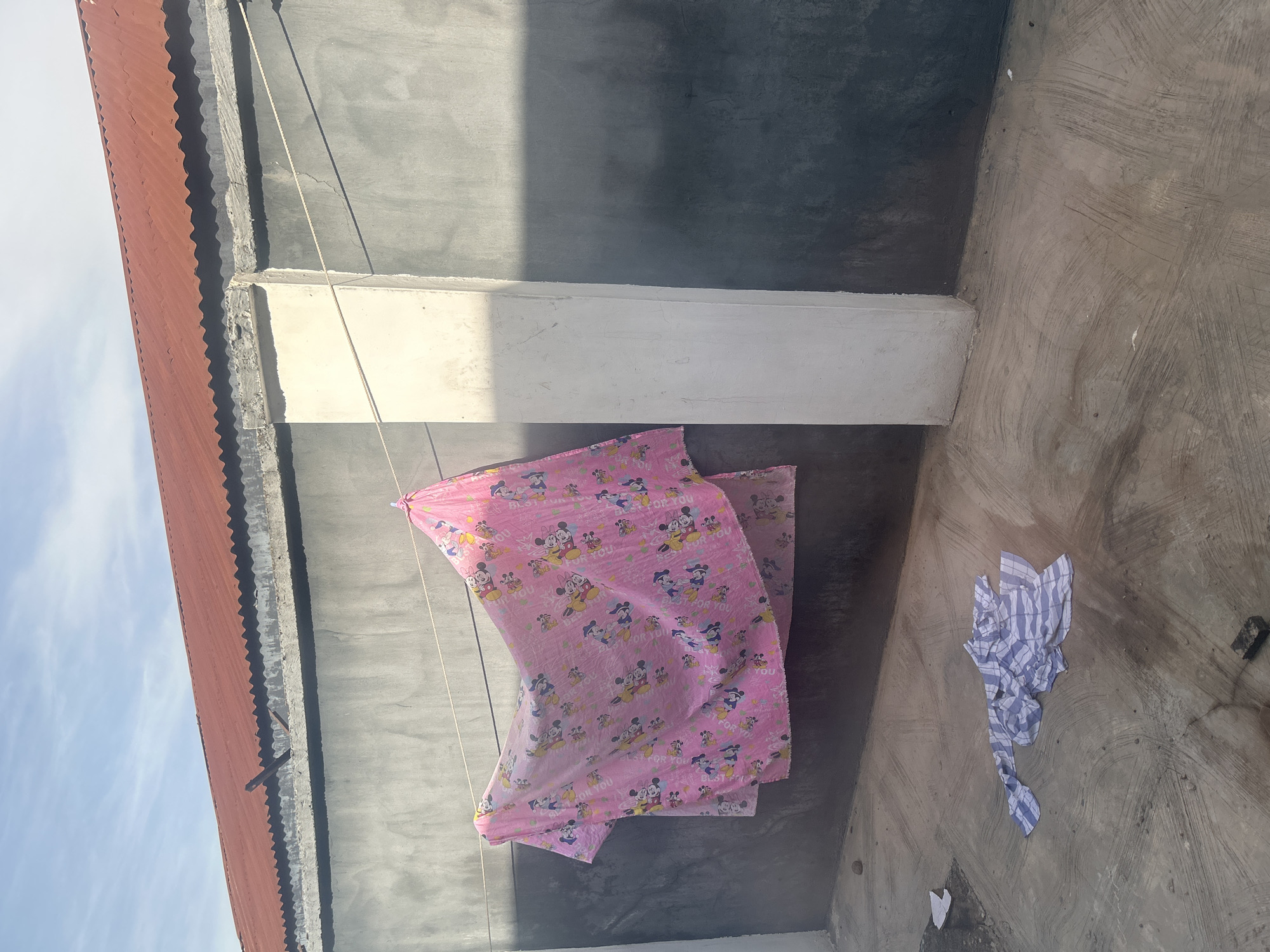 The image depicts a bright, outdoor scene, likely a residential backyard, in Ogbomosho, Nigeria. The foreground is a concrete ground with a pattern of lines suggesting a brushed finish. A crumpled pile of blue and white striped fabric lies on the ground to the left, with some scattered white paper fragments nearby. The midground is dominated by a wall with a rough, gray plastered surface. A clothesline stretches horizontally across the frame, supporting a large pink fabric with a pattern of cartoon characters, possibly Disney's Mickey Mouse. The fabric is attached to the line with a white clothespin visible on the right. To the right of the fabric, a tall, white rectangular pillar juts out from the wall, casting a distinct shadow. The background shows the top of the wall, with a corrugated red metal roof extending above it, angled downwards. The sky above is a clear blue with a few wispy clouds. The lighting suggests it is daytime, likely late morning or early afternoon, with sunlight casting strong shadows.