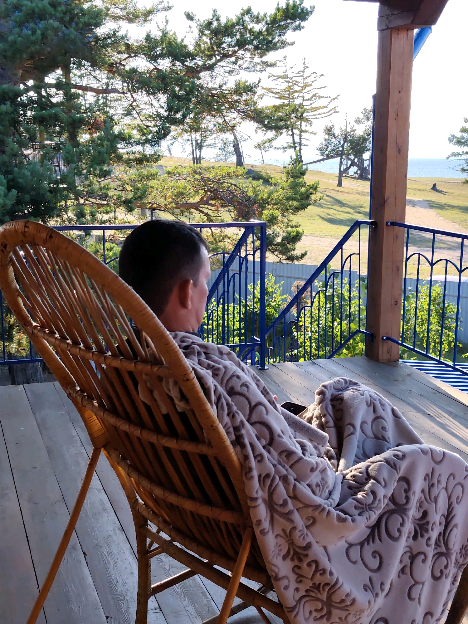 A man, viewed from behind and from the right, sits in a woven wicker chair on a wooden porch. He is wrapped in a light-colored blanket with a patterned design, and he holds a dark-colored smartphone in his right hand. The man has short, dark hair and is wearing a dark shirt. The porch has a wooden floor and a railing with blue metal decorative elements and blue metal posts. In the background, there is a grassy slope with scattered trees, and beyond that, a large body of water, likely Lake Baikal, under a bright, hazy sky. The scene appears to be in Kharantsy, Russia, characterized by its natural landscape and a structure with a porch overlooking the lake.