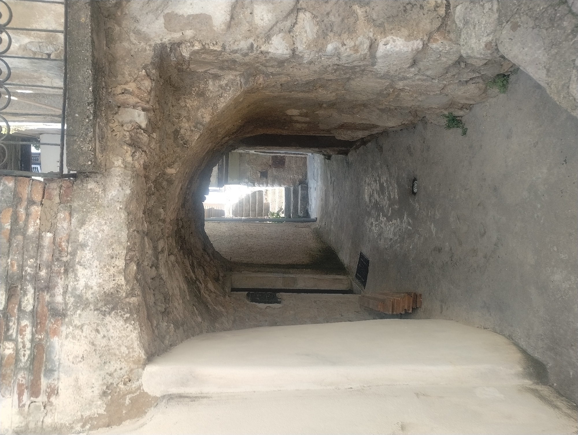 An outdoor, ground-level view captures a narrow, arched passageway constructed from rough stone and mortar, with a worn concrete floor. To the left, a smooth, cream-colored wall curves outward, showing some dark staining near the bottom. To the right, the passageway is defined by a rugged stone wall that follows its curve. The archway itself is composed of rough-hewn stones and appears weathered.
Beyond the arch, the passageway opens to a view of stone steps ascending towards an area bathed in sunlight. A dark, rectangular doormat lies on the concrete floor near the entrance to a dark doorway set into the stone wall on the left. A stack of wooden planks is visible next to this doorway. A small, dark metal grate is embedded in the concrete floor within the archway. Some sparse green vegetation grows from the cracks in the stone wall on the right, near the end of the passageway. The scene suggests a historic, possibly Mediterranean, urban setting. Based on the architectural style and the information provided, this location is likely within the town of Vrbnik on the island of Krk, in the Primorje-Gorski Kotar County of Croatia, a region known for its stone architecture and narrow alleyways.
