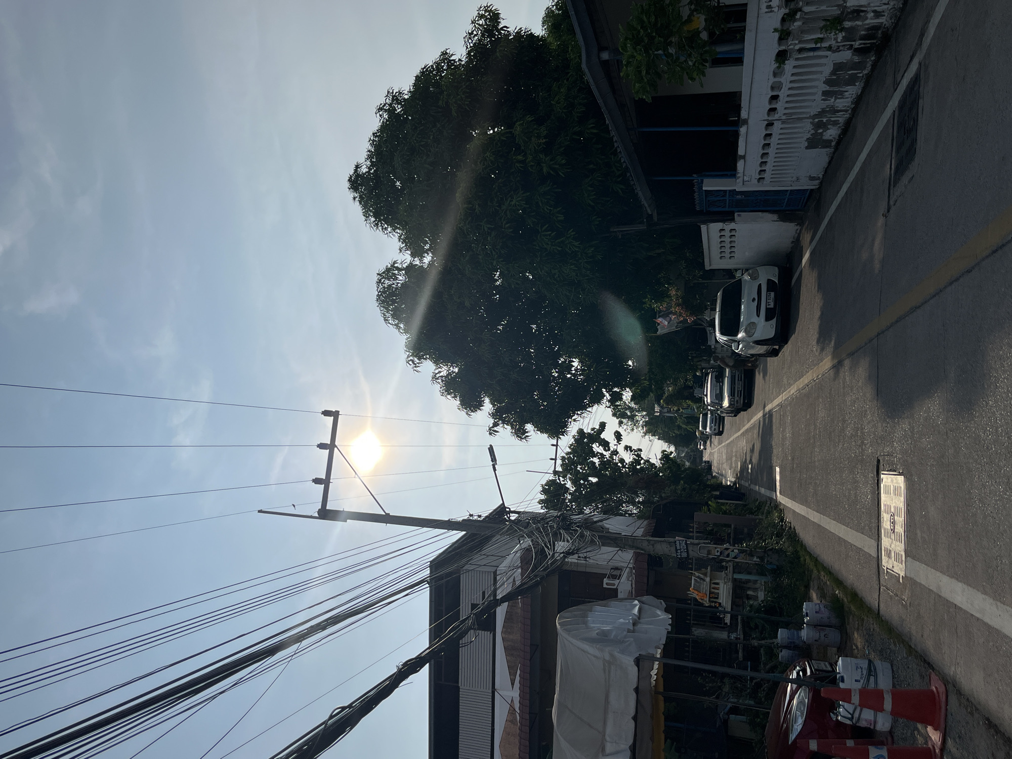The image depicts a street scene in Pattaya City, Thailand. The view is along a narrow road with buildings on either side. The sky is light blue with a bright sun. Power lines are visible in the foreground. On the right side of the road, there are parked cars. There are several paint cans on the ground near the front of a red vehicle.
