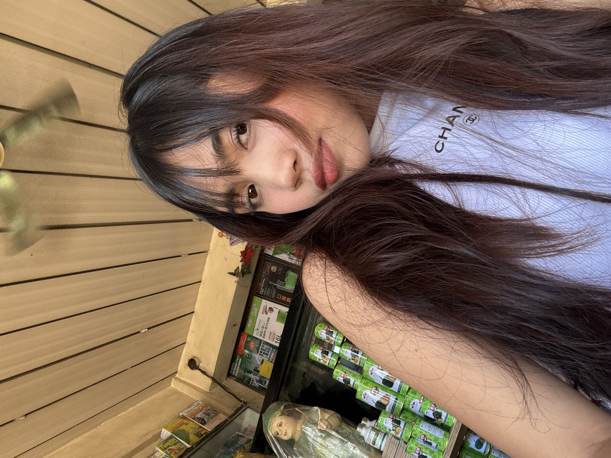 This is a close-up, eye-level selfie of a young woman in Tagum City, Philippines. She has long, dark brown hair with bangs that partially obscure her face. She is wearing a white t-shirt with a black Chanel logo visible on the chest. The background suggests she is in a store or market stall. Shelves with various products are visible, including green cans stacked neatly, some with cartoonish illustrations. To the left, there is a veiled statue of a child-like figure, possibly a religious icon. The ceiling above is made of light-colored wooden planks. A ceiling fan is visible in the upper portion of the image, indicating an indoor setting. The lighting is warm and appears to be natural sunlight.
