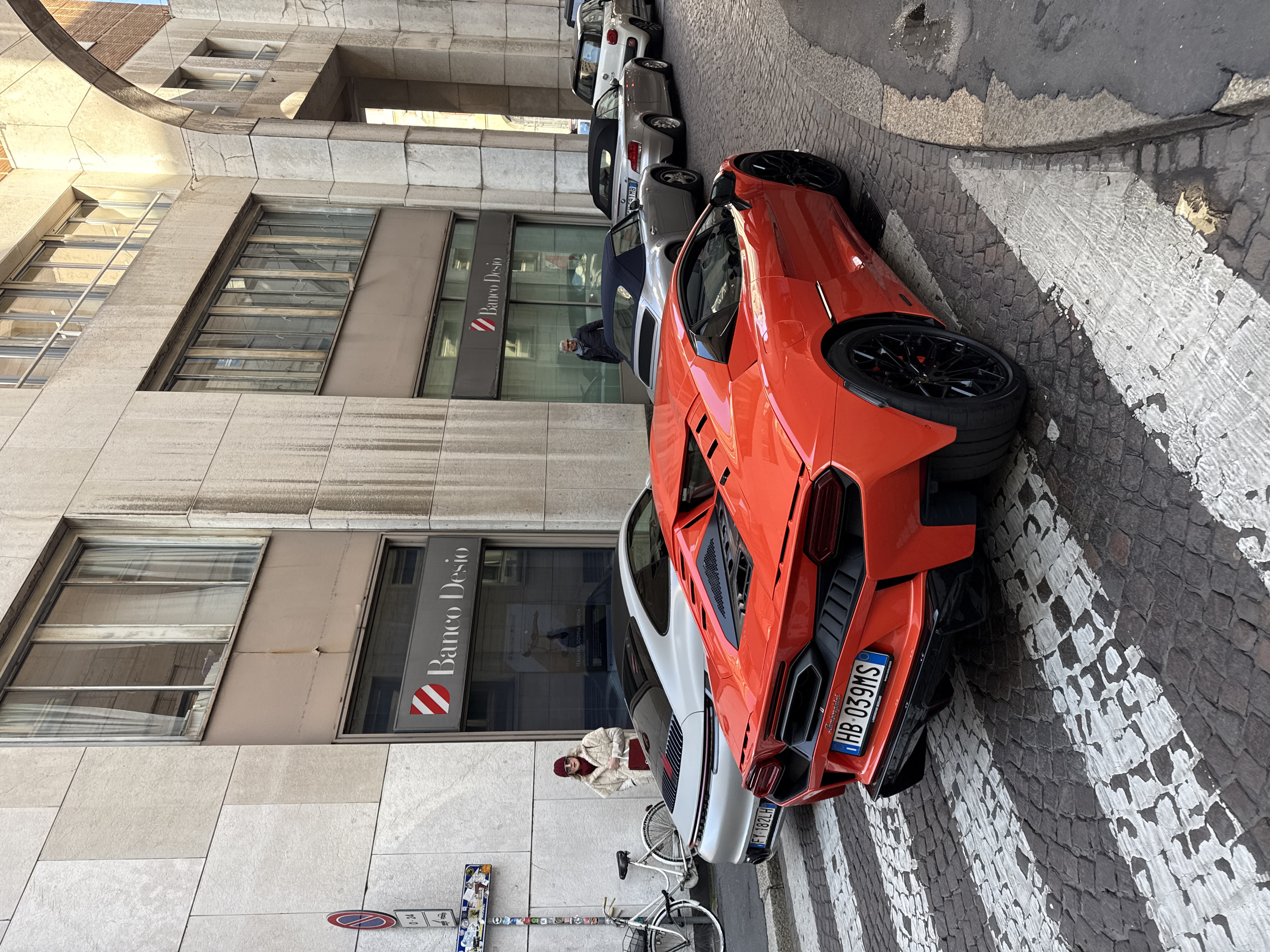 The image depicts a street scene in Milan, Italy, characterized by urban architecture and luxury vehicles. In the immediate foreground, an orange Lamborghini, identified by its rear badging and Italian license plate "HB 039MS," is parked facing away from the viewer, partially on a white-striped pedestrian crossing over cobblestone paving. Its rear features dark hexagonal exhaust vents, horizontal taillights, and black multi-spoke wheels.
Behind the Lamborghini, a silver Porsche, with an Italian license plate "FY 182LH," is parked. A white bicycle leans against the building facade to the left of the Porsche. Further along the street to the right, a line of parked cars includes a silver BMW Z4 convertible with its top down (partially visible license plate "DW 291MB") and a white Fiat 500.
Two individuals are present on the sidewalk. On the left, a woman wearing a cream-colored faux fur coat, a dark red beanie, a matching scarf, and sunglasses, holds a dark red handbag. She stands near the entrance of "Banco Desio." Further down the sidewalk to the right, a man with grey hair, dressed in a dark jacket and blue scarf, stands near another section of the same bank.
The prominent building on the left and center features a light-colored paneled facade with multiple windows, some reflecting the sky or opposite buildings. The facade exhibits vertical streaking, indicative of weathering. The bank "Banco Desio" has its logo (a red and white striped shield) and name displayed at street level. On the far right, a large, light-colored stone archway extends across the street or connects building sections. A street pole on the left sidewalk displays a "No Parking" sign with a "0-24" duration and is covered in various stickers. The street is paved with weathered cobblestones, and the white pedestrian crossing stripes show signs of wear. The scene is illuminated by natural daylight.