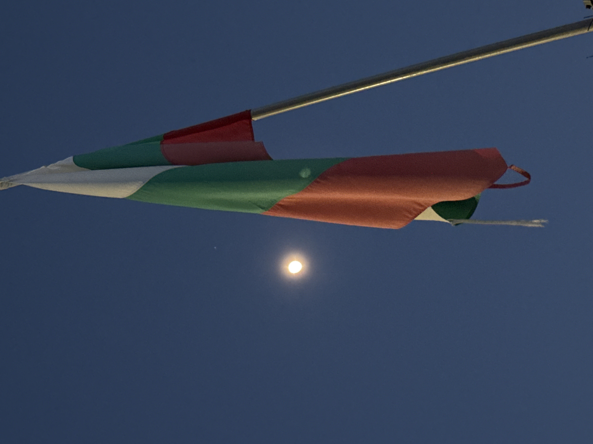 The image shows a Bulgarian flag on a metal pole against a dark blue sky in Galishte, Bulgaria. A bright, spherical light source is visible in the background.