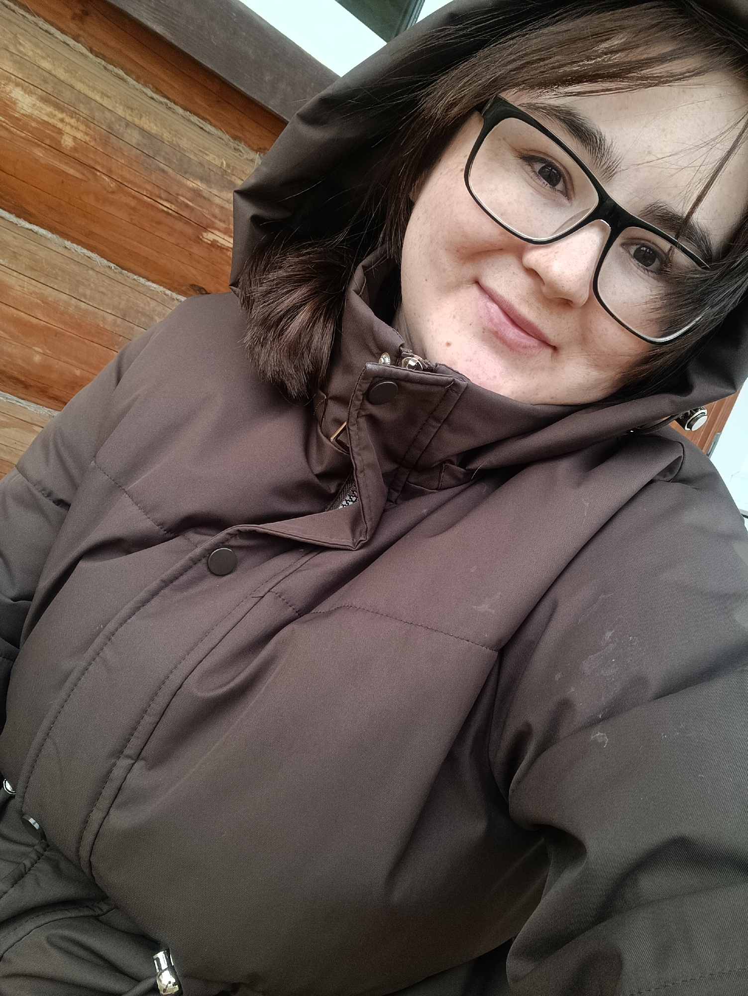 A close-up selfie depicts a young adult, likely female, with dark brown hair and black-framed glasses, smiling gently at the camera. Her face features freckles and a pleasant expression. She is wearing a dark brown or charcoal-colored puffer jacket with the hood up. The jacket has visible stitching, dark buttons, and metallic toggles on drawstrings around the neck and waist.

The background primarily consists of a rustic wooden log wall, showcasing horizontal logs with a reddish-brown tone, suggesting a traditional building or cabin. A bright, indistinct area is visible in the upper right, possibly indicating an outdoor setting or a window. The attire suggests cool weather, perhaps autumn or winter. The overall scene, with the wooden architecture, could be characteristic of a rural or suburban environment in a location like Barda, Russia. No visible text is present in the image.