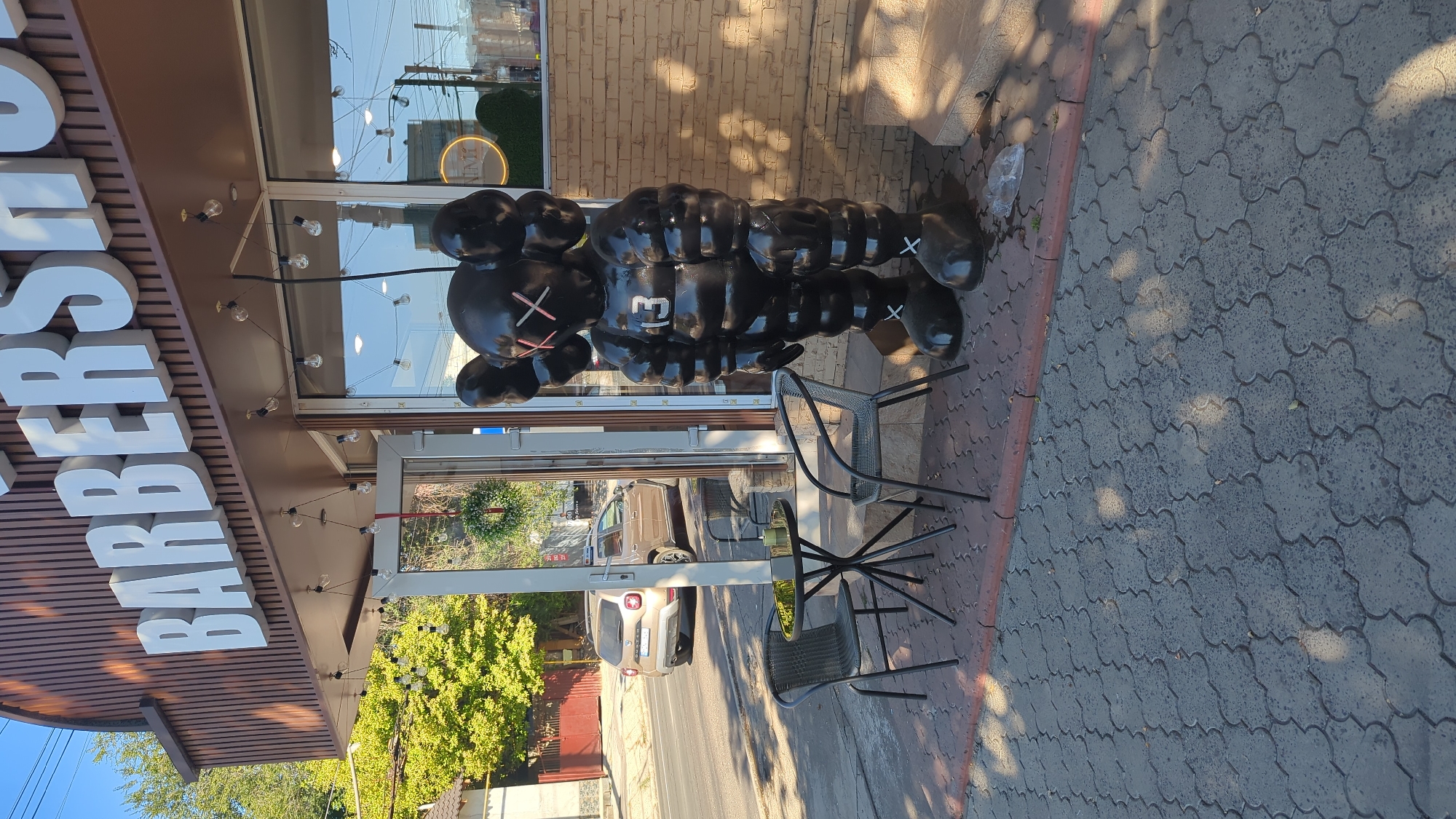 The image shows a street view in Chișinău, Moldova, featuring the exterior of a barbershop. The barbershop has large white lettering spelling "BARBERSHOP" on its facade, and the building is adorned with string lights. In front of the shop, there is a black, inflatable-style statue of a cartoonish figure with X's for eyes and the number "13" on its chest. A small round table with two chairs sits on the sidewalk in front of the statue. A tan car is parked on the street to the left of the scene, and trees and a building are visible in the background. The ground is paved with interlocking concrete blocks.