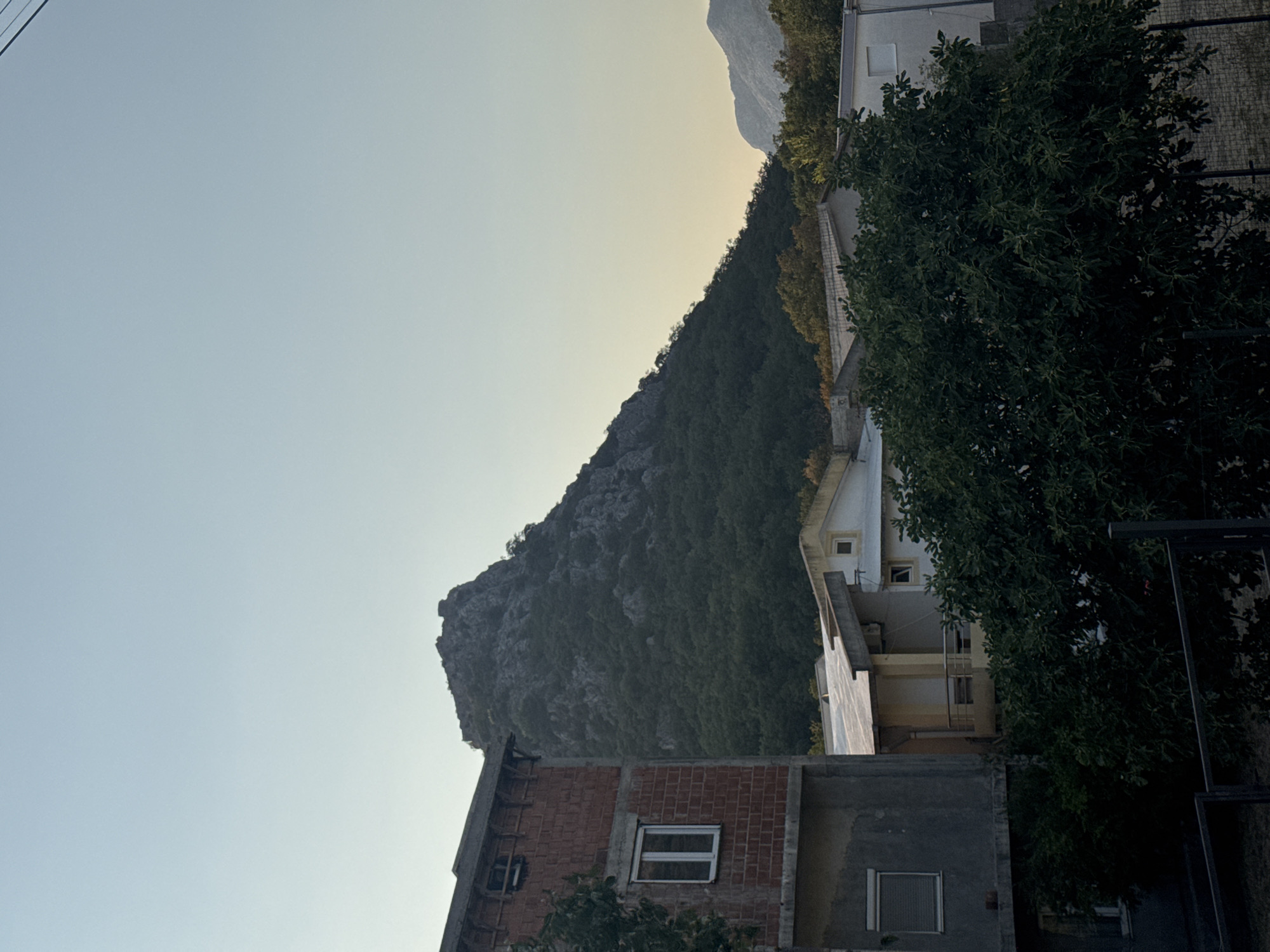 In Sutomore, Montenegro, a view unfolds with a stark, unfinished brick building in the foreground, its concrete foundation and upper levels exposed. To its right, a cluster of houses with pitched roofs and white facades line the landscape. Dominating the background is a rugged, tree-covered mountain, its peak jagged and rocky. The sky above is a pale, clear blue, transitioning to a warm yellow hue near the horizon, suggesting either sunrise or sunset. The scene is quiet, with no people or discernible activity, presenting a tranquil, somewhat austere view of the Montenegrin coast.
