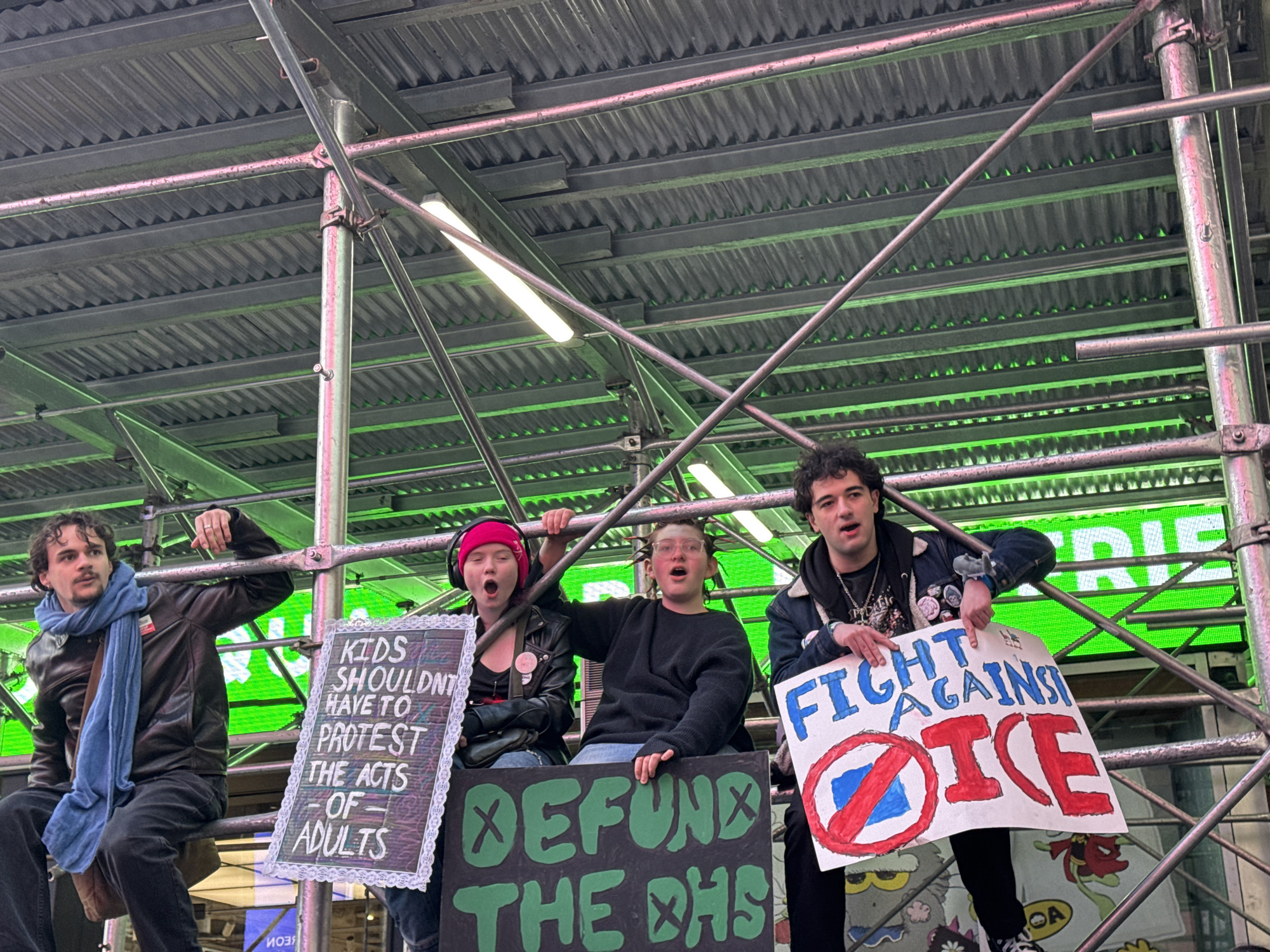 Four people are sitting on scaffolding in New York City, holding protest signs. The person on the left is a man with a mustache and blue scarf, sitting with his legs dangling. Next to him is a person with a pink hat, mouth open, holding a sign that reads "Kids shouldn't have to protest the acts of adults". The next person has their mouth open and is holding a sign that reads "Defund the DHS". The person on the right is holding a sign that reads "Fight against ICE". The background consists of scaffolding, fluorescent lights, and a large digital screen displaying green text. The environment is urban.