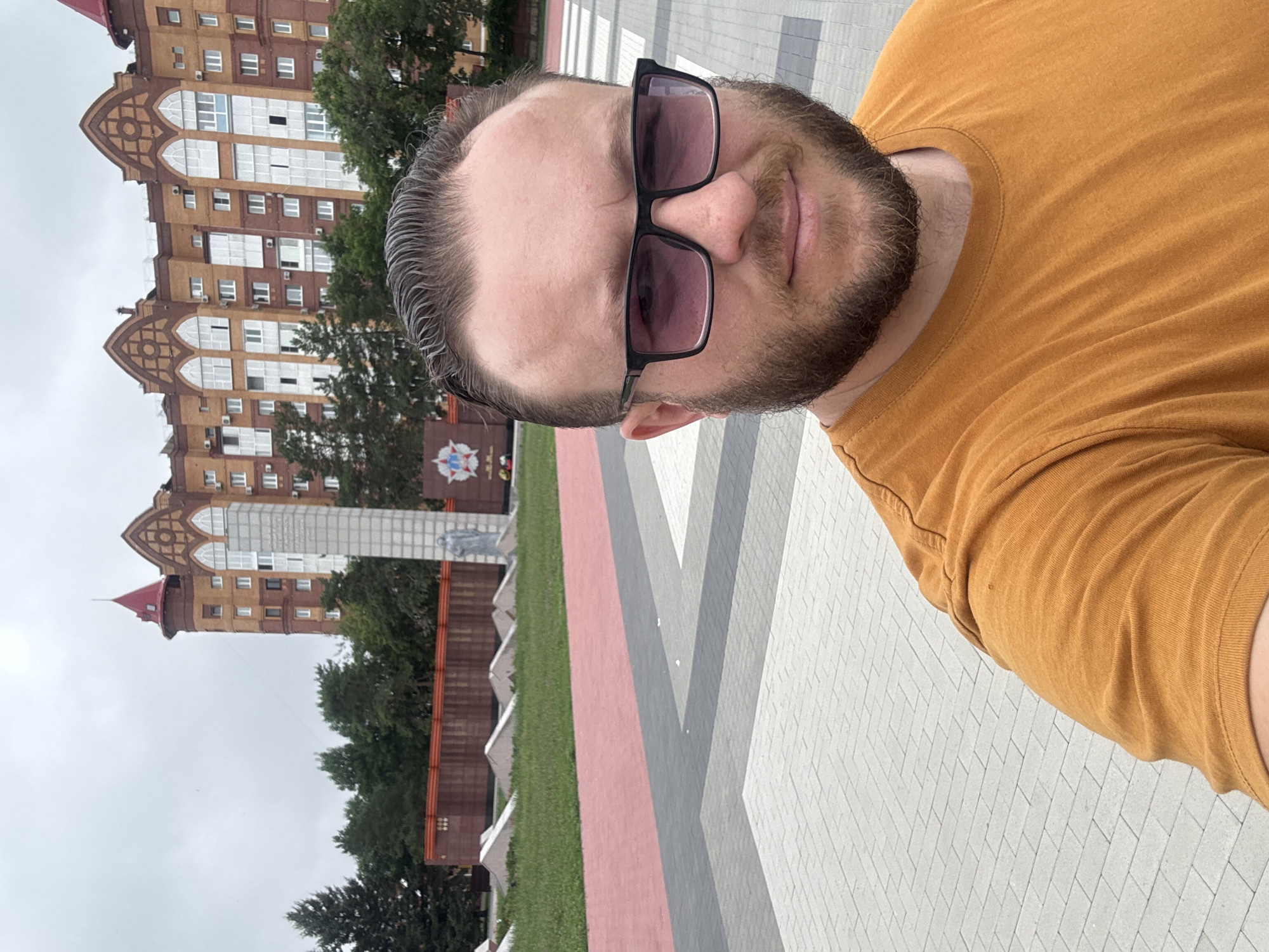 The image is a selfie taken outdoors in the city of Blagoveshchensk, Russia. In the foreground, a man with a beard and sunglasses is visible, wearing an orange t-shirt. He is standing on a paved walkway with a geometric pattern. In the background, there is a war memorial with a tall obelisk and a statue. Behind the memorial, a modern residential building with an orange and brown facade and a red turreted roof is visible. Trees and a grey, overcast sky complete the scene.