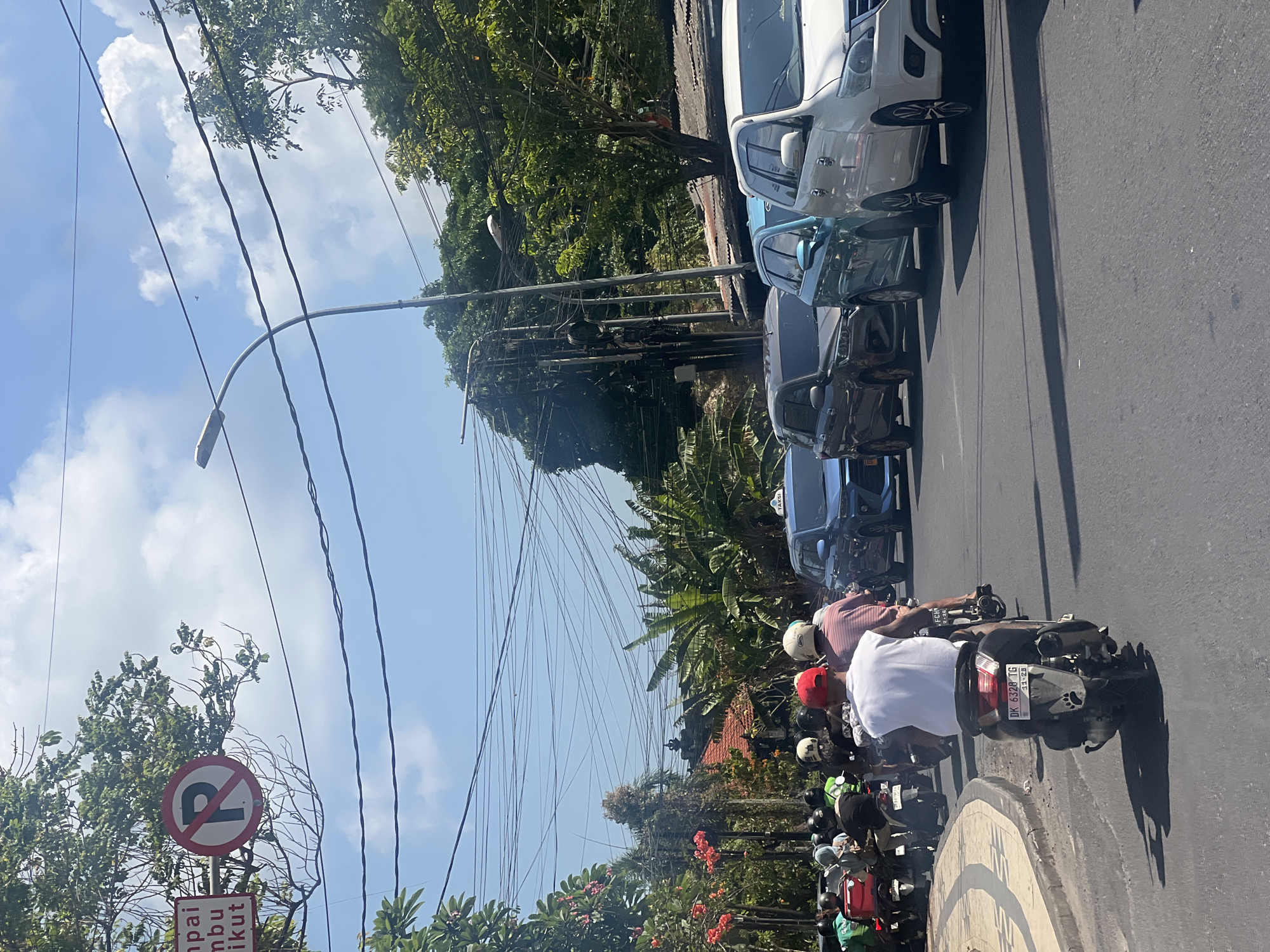 The image depicts a street scene in Kuta, Indonesia, likely during daylight hours given the bright sunlight and clear sky with scattered clouds. A line of vehicles, predominantly motorcycles and several cars, are stopped or moving slowly in traffic on an asphalt road.
In the foreground, a person wearing a white sleeveless shirt and a red baseball cap is riding a dark-colored scooter. The scooter has a license plate that reads "DK 6328 TG". Behind this scooter, other motorcycles are visible, many with riders wearing helmets. To the right of the scooters, a line of cars is present, including a white SUV, a teal SUV, and a dark gray SUV. The white SUV has "TAXI" written on its roof.
The environment shows lush green trees and foliage on both sides of the road, with some red flowers blooming on the left. Overhead, a complex network of power lines crisscrosses the sky, supported by poles. A street light is also visible on the right. On the left side of the road, a circular sign with a red border and a red diagonal line over a black 'P' symbol indicates "no parking". Another rectangular sign with text, partially obscured by foliage, is also present. The scene suggests a typical urban traffic situation in a tropical climate.
