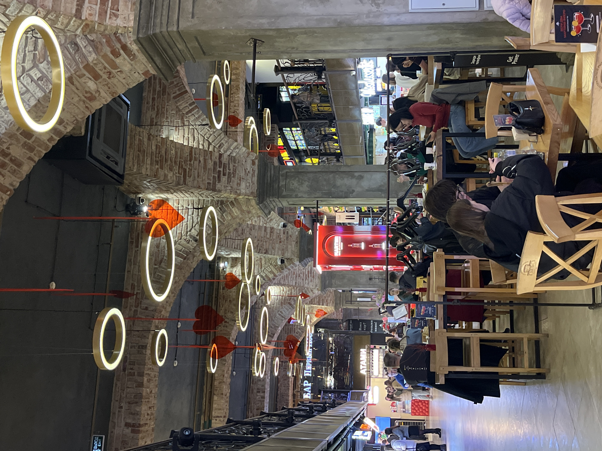 The image captures the interior of a restaurant or bar, possibly located in the city of Moscow, Russia, given the Cyrillic script present on signage. The location has an industrial aesthetic, with exposed brick arches adorned with hanging circular lights, and red heart-shaped decorations suspended from the ceiling. Patrons are seated at wooden tables, some with drinks or menus visible. The scene appears to be bustling with activity, as people are conversing and engaging with their mobile devices. A sign displays "БАР ПЕРВЫЙ АТАМИ," which translates to "First Bar Atoms." Another sign reads "Заряди телефон Get Power," indicating a charging station for mobile phones. Additional signage includes "MARTINI." The overall atmosphere suggests a contemporary and lively environment, potentially catering to a diverse clientele.