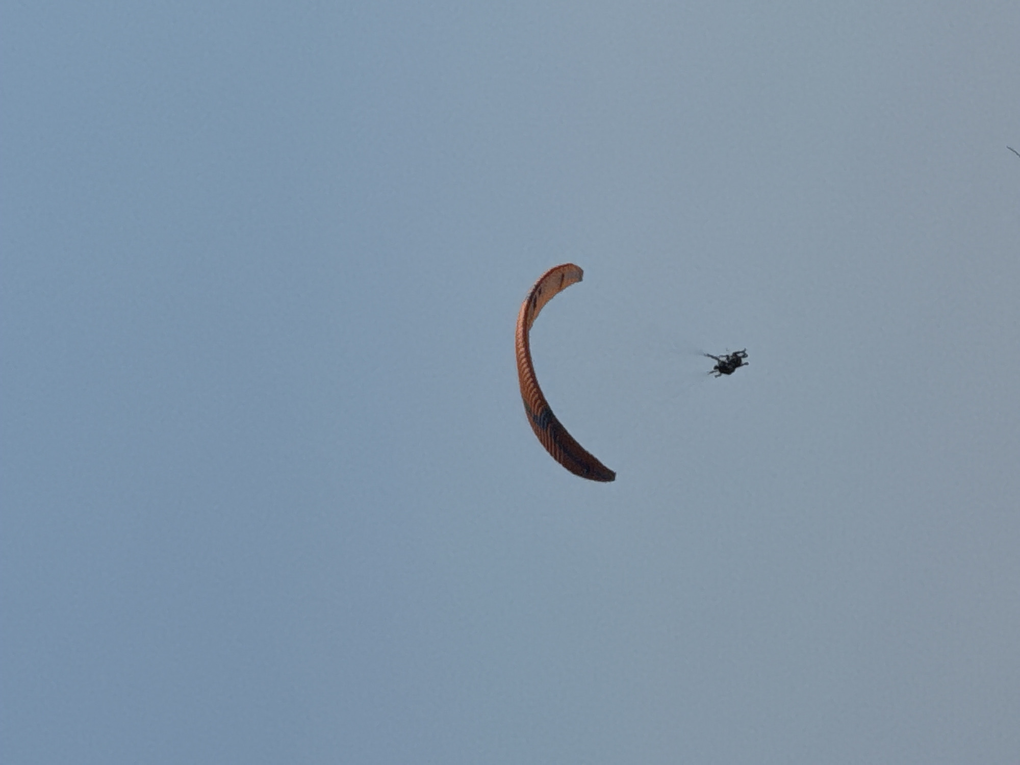 A paraglider with two individuals onboard is captured against a clear, pale blue sky. The paraglider's canopy is a vibrant orange with dark blue accents, arching prominently in the upper middle of the frame. Below the canopy, two figures are suspended in a harness, appearing to be a tandem paragliding experience. The scene is set against an expansive, featureless sky, indicating an outdoor activity in an open area, likely a scenic overlook or a location popular for airborne sports. The image does not provide specific geographic markers to confirm a location within Boreti, Montenegro, but the general environment suggests a mountainous or coastal region given the activity.