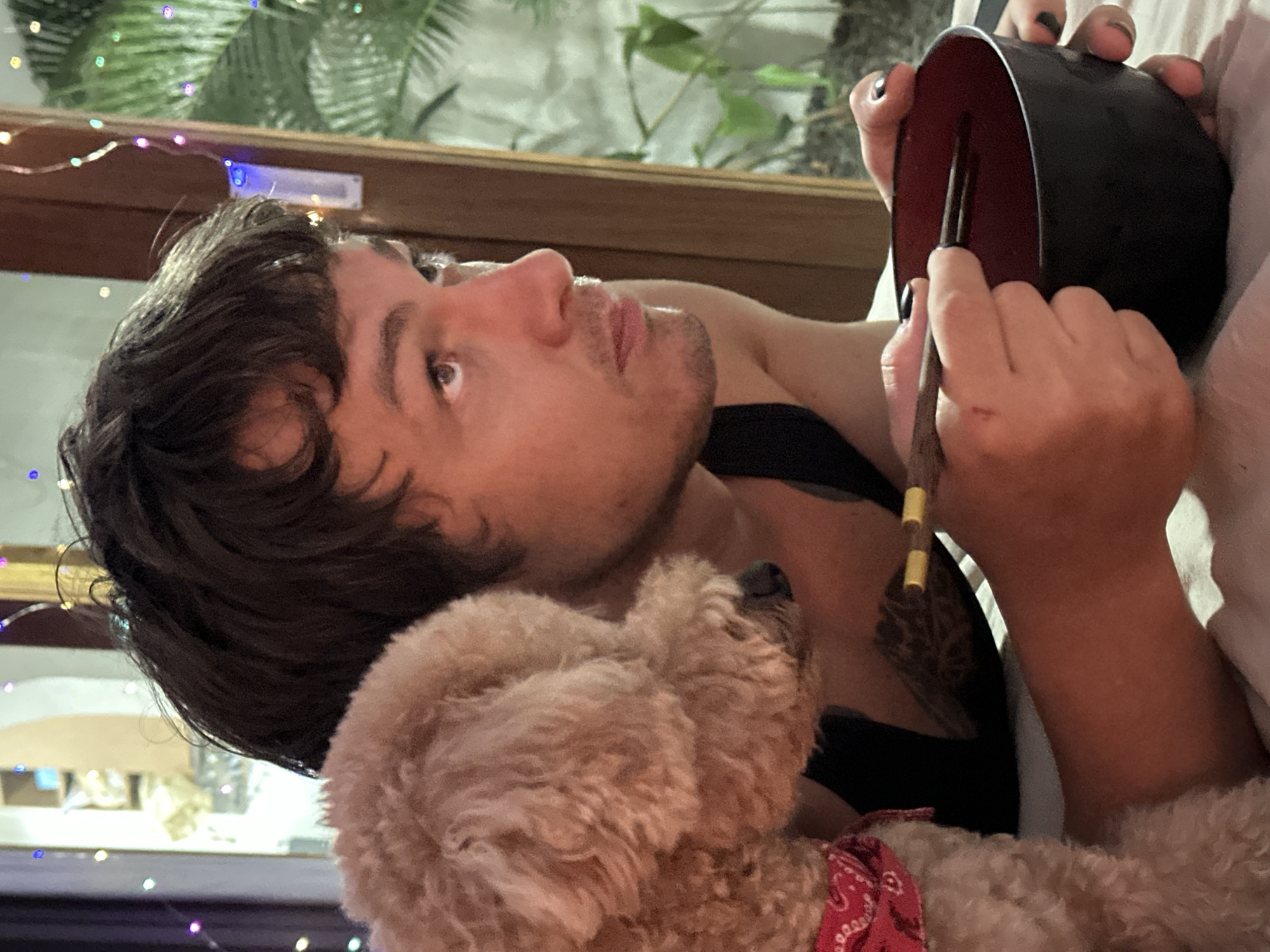 A close-up shot depicts a man lying on his side, possibly in an indoor setting, with a dog nestled close to him. The man has dark hair and light skin, his face turned upwards and to the side. He appears to be holding a black bowl with a red interior, and chopsticks rest inside it. The man's fingernails are painted black. The dog, a small, furry breed, is positioned near the man's neck, wearing a red and white bandana. The man has a tattoo visible on his upper chest, next to a black tank top. The location is North Kuta, Indonesia. The environment features greenery visible through a window in the background, along with some decorative string lights. The scene suggests a relaxed, domestic atmosphere.