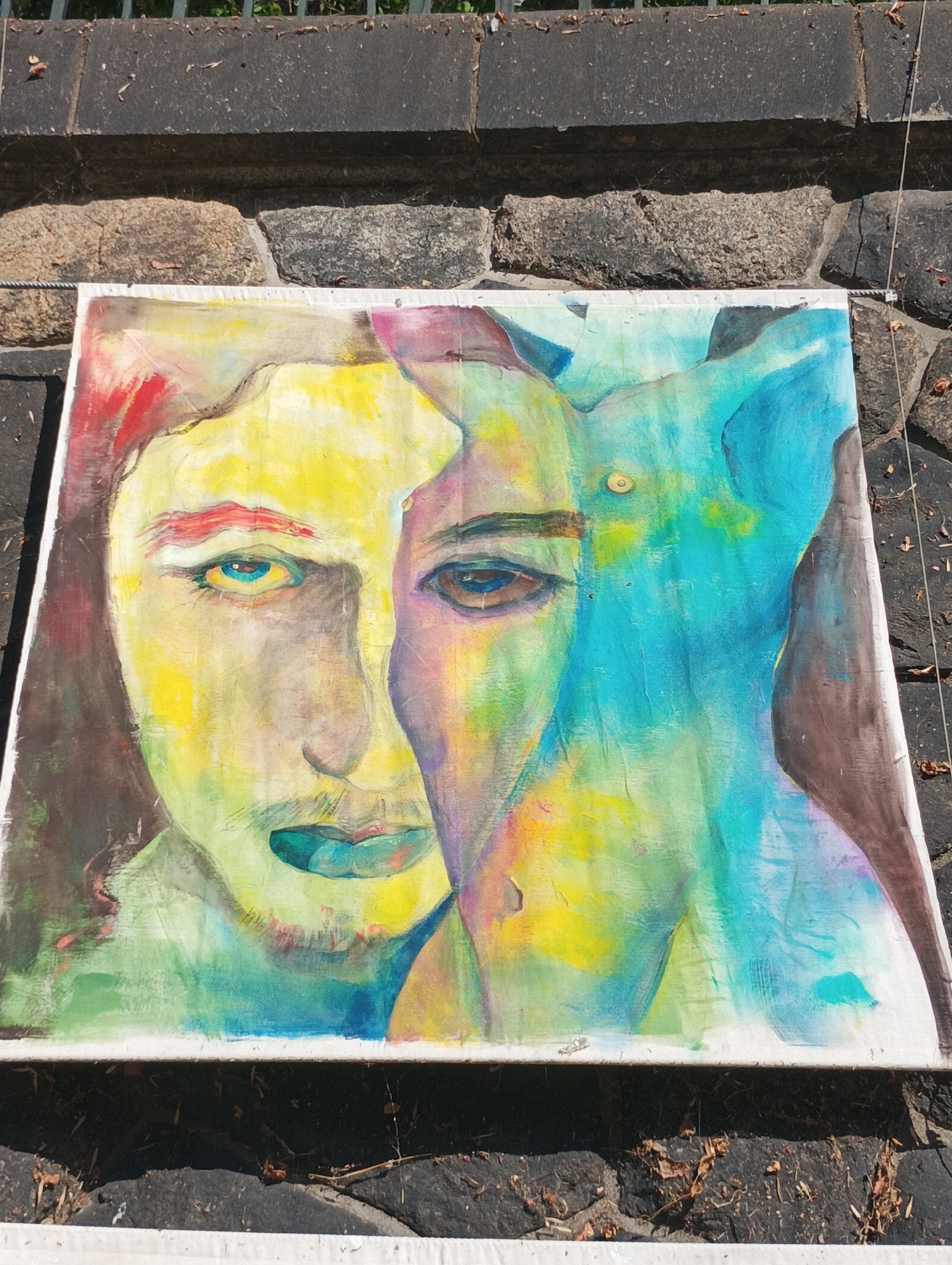 A large, colorful abstract painting featuring two overlapping faces is displayed outdoors, possibly as part of an exhibition or street art. The painting is positioned horizontally and appears to be on a canvas. One face is predominantly yellow with red accents on the eyebrows and a blue-green mouth. The other face, partially obscured, shows darker hues and brown eyes. Vibrant blues and greens dominate the background and blend into the faces, creating a surreal and dreamlike effect. The painting is propped up against a textured stone wall, with a metal railing or wire visible above it. The ground in front of the painting is paved with dark stones and littered with dry leaves and debris. The overall setting suggests an outdoor urban environment in Pilsen, Czechia, where art is publicly displayed.