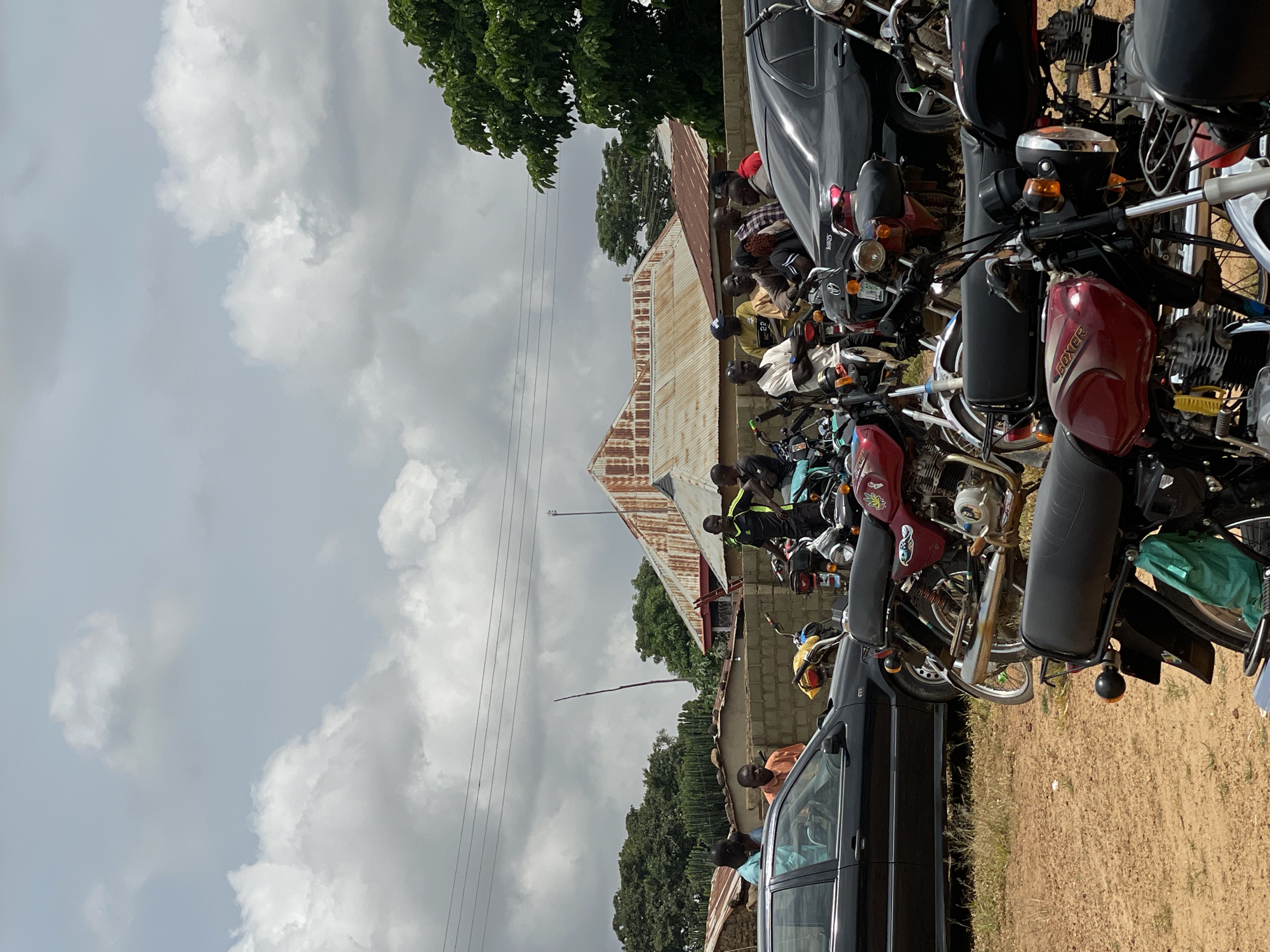 The image depicts a street scene in Kassa, Nigeria, under a cloudy sky. A group of men are gathered around several motorcycles and two black cars parked on a dirt road. The men, dressed in casual attire, are standing and talking. The motorcycles are positioned in the foreground and middle ground, with some of the men sitting on them. In the background, there are buildings with corrugated metal roofs and a few trees. Power lines are visible across the sky. The overall impression is of a casual gathering in a semi-urban environment.