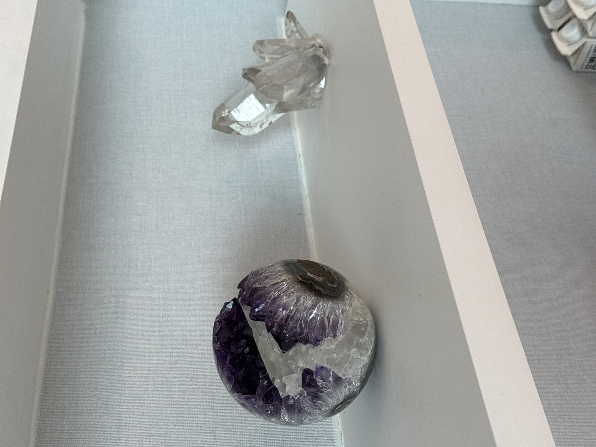 The image shows a white drawer containing two decorative stone pieces. One is a cluster of clear quartz crystals, and the other is a geode sphere, purple and white. The location is unknown, and there's no indication that this image was taken in Miami, Florida. There are no people in the image.