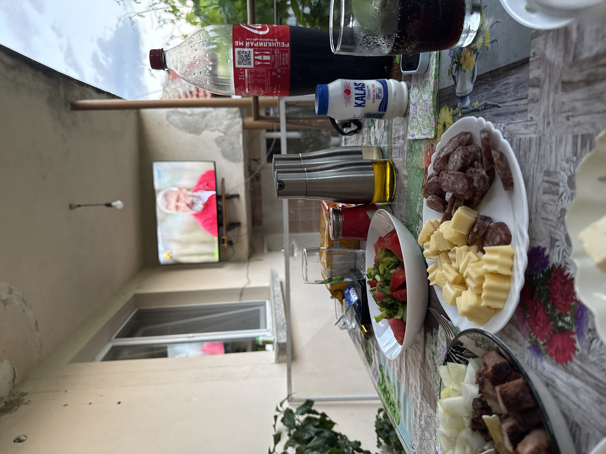 Here's a detailed description of the image:
**Setting:** The scene appears to be an outdoor meal setting on a patio or similar outdoor space. A TV is visible in the background showing a person with a white beard and red clothing.
**Objects:**
* **Food and Drink:** There's a table laden with various food and drink items. These include a bottle of Coca-Cola, glasses of dark liquid (possibly more Coca-Cola), a bottle of salt ("KALAS Classic Iodized Sea Salt"), a metal bottle likely containing oil or vinegar, a plate with cheese and cured meat slices, a bowl of salad (tomatoes and lettuce are visible), and a dish with what seems to be grilled meat and chopped onions.
* **Table Setting:** The table has a patterned tablecloth. Cutlery is also present.
* **Background:** In the background there is a TV screen and some construction/repair activity on a building.
**Location:** The Coca-Cola bottle is labeled with Bulgarian text "РЕЦИКЛИРАЙ МЕ ЗА РЕЦИКЛИРАНЕ СЪМ". Also, the bottle of salt is labeled "KALAS". Based on this and your prompt I am going to say that the location is Sarnevo, Bulgaria.
**Overall Impression:** The image captures a casual outdoor meal setting, possibly a gathering with friends or family. The presence of the TV suggests people may be enjoying food while watching a show.