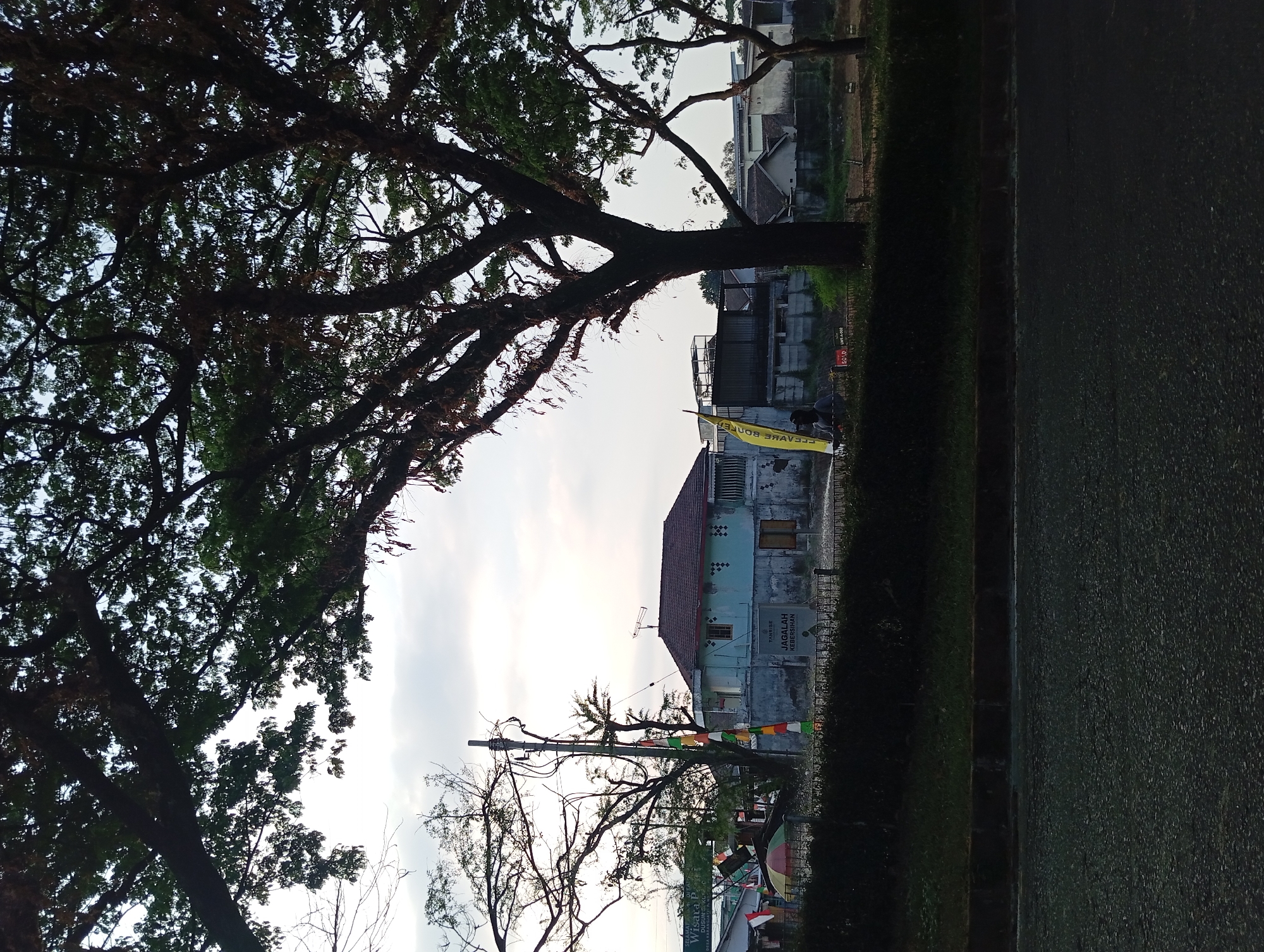 The image captures a street scene in Ketanireng, Indonesia, under a cloudy sky. In the foreground, a dark asphalt road is visible, bordered by a strip of grass and then a dense green hedge. Behind the hedge, a light blue house with a brown tiled roof stands prominently. The house has several windows and a weathered facade. To the left of the house, a utility pole with electrical wires is present. A few colorful flags are attached to the pole and a nearby fence. A yellow banner with black text is also visible, leaning against a tree. In the middle ground, various buildings, including residential structures and what appear to be commercial or industrial buildings with metal roofs, are situated behind the house and hedge. Large trees with full canopies dominate the upper portion of the frame, casting shadows and framing the scene. A small, colorful umbrella is partially visible behind the hedge on the left. The overall impression is of a quiet, possibly rural or semi-urban, residential area.