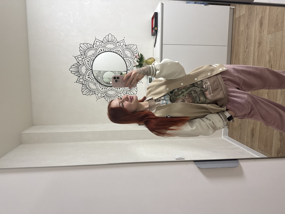 A person with red hair takes a selfie in a mirror. The reflection shows a woman wearing a tan jacket, graphic t-shirt, and pink pants. Behind her, a mandala design surrounds a circular mirror on a white wall. A white cabinet with black accents is visible in the background, along with light wood flooring. The image is taken from a slightly low angle and the mirror's frame is evident at the bottom.