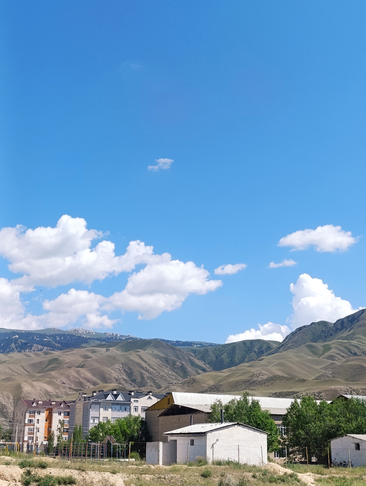 In the city of Naryn, Kyrgyzstan, a clear blue sky with scattered white clouds stretches above a landscape of rolling, barren hills. In the foreground, a cluster of apartment buildings, painted in various shades of beige, orange, and gray, are situated against the backdrop of these hills. Trees line the street in front of the buildings. To the right, a long, white-roofed building and several smaller, simple structures with white walls and corrugated metal roofs are visible. A chain-link fence encloses a small playground area with metal equipment. The overall scene depicts a semi-urban residential area nestled within a mountainous, semi-arid environment.