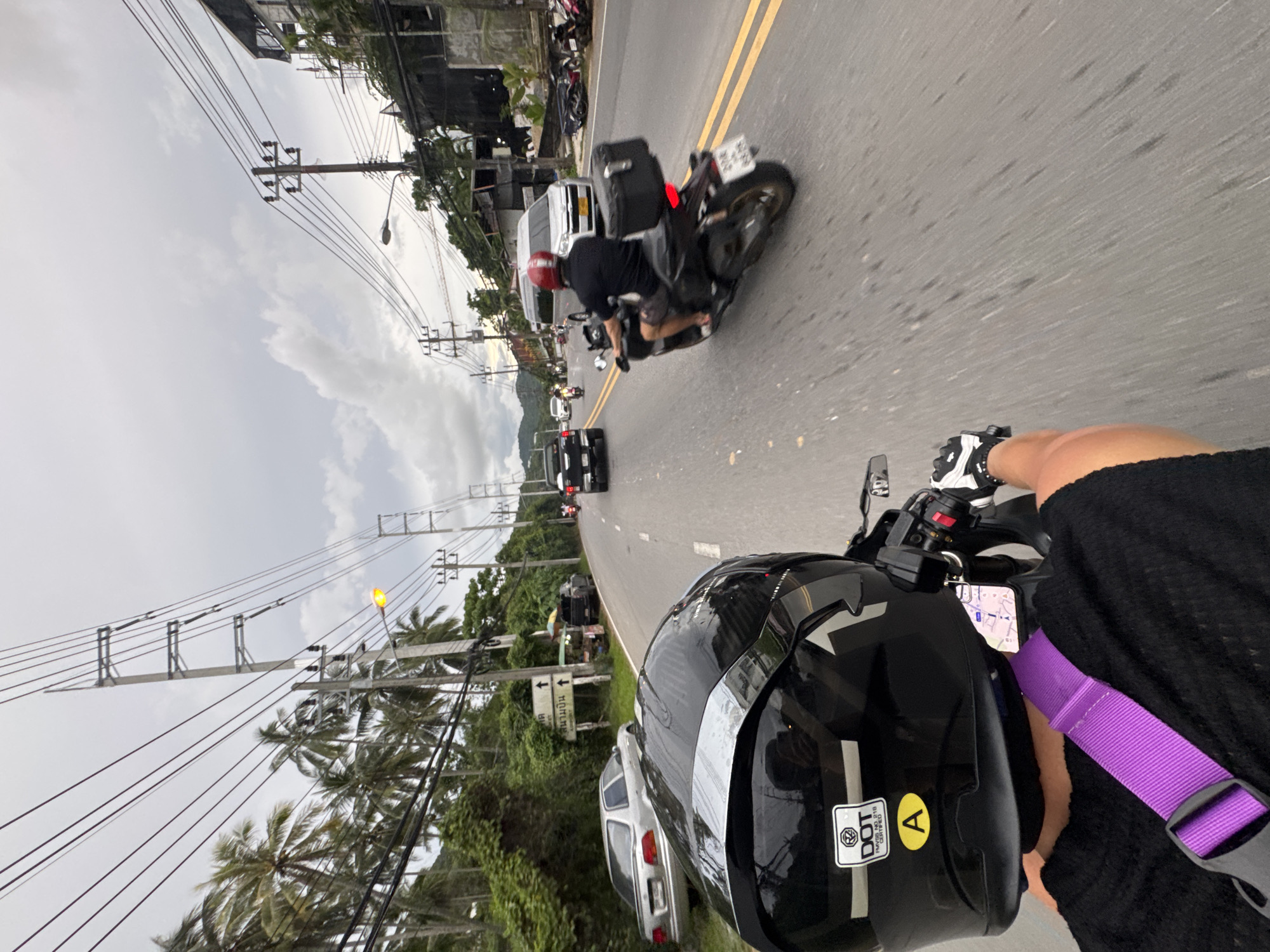 The image shows a first-person perspective of someone riding a motorcycle in Karon, Thailand. The road is paved with yellow lane markings and lined with palm trees and vegetation on the left. On the right side of the road, there are buildings and more power lines. Several vehicles are visible, including cars, vans, and other motorcycles. In the foreground, the rider's black helmet with a yellow "A" sticker is prominent. The rider is wearing a black shirt and a purple strap across their chest, and white and black gloves. In the middle ground, another person is riding a dark-colored scooter, wearing a black shirt and a red helmet, with a black storage box on the back. The sky is overcast with clouds. There are also street signs and a small food stall visible along the side of the road.