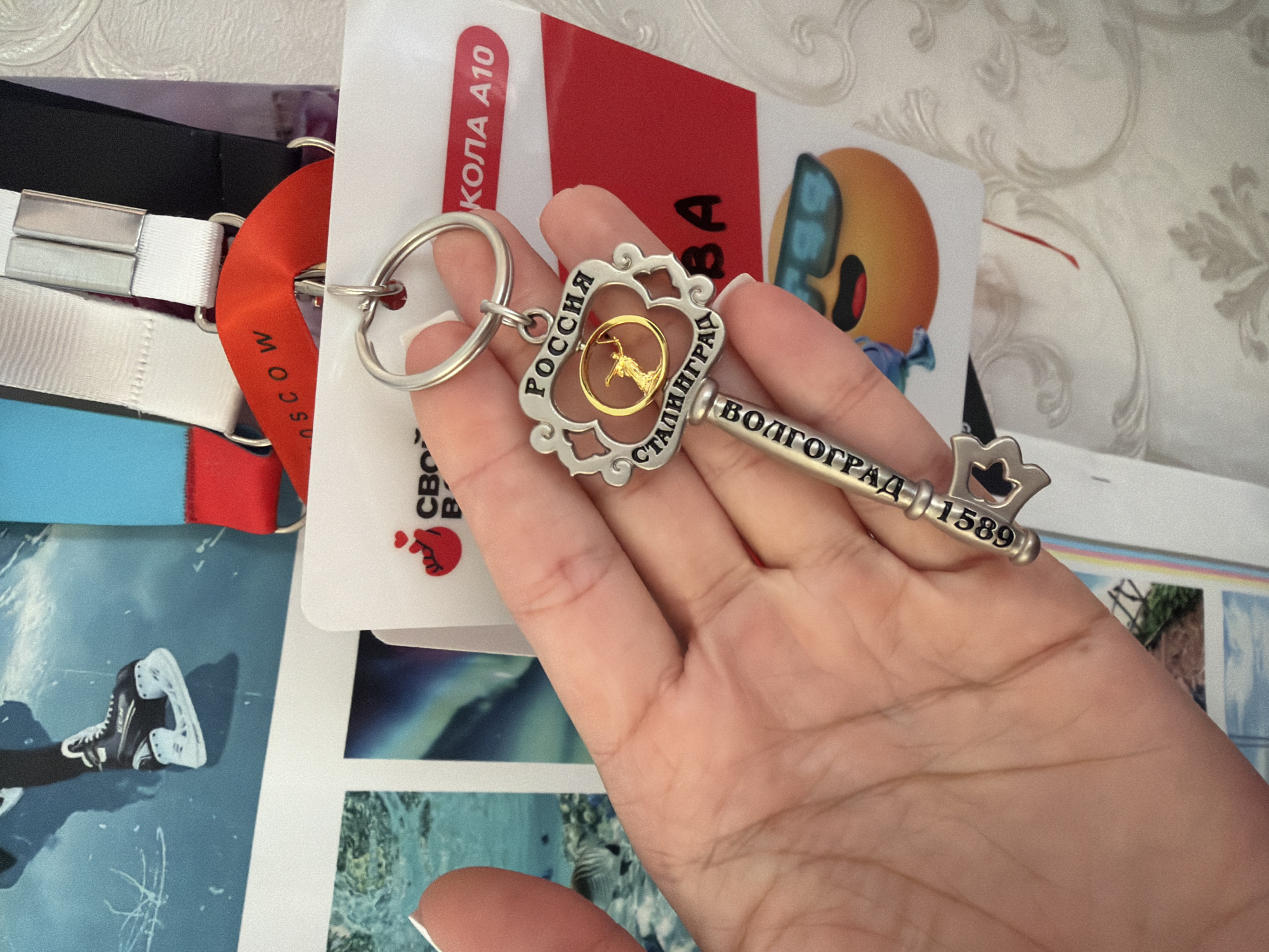 In the foreground, a person's hand holds a decorative key keychain. The keychain has an ornate design and features the text "РОССИЯ" (Russia) and "СТАЛИНГРАД" (Stalingrad). Below this, the key bit has the text "ВОЛГОГРАД 1589" (Volgograd 1589). The keychain is attached to a silver ring. The hand is positioned in front of several items, including a white card with text that appears to be in Russian and a red ribbon with the word "MOSCOW" visible. In the background, there are other items, including what appears to be a picture of a figure skating boot and various colored lanyards. There is also a sticker of an emoji wearing sunglasses. The overall setting appears to be indoors, possibly a home or office, given the presence of what looks like patterned wallpaper. There is no clear indication of the location being in Sochi, Russia.