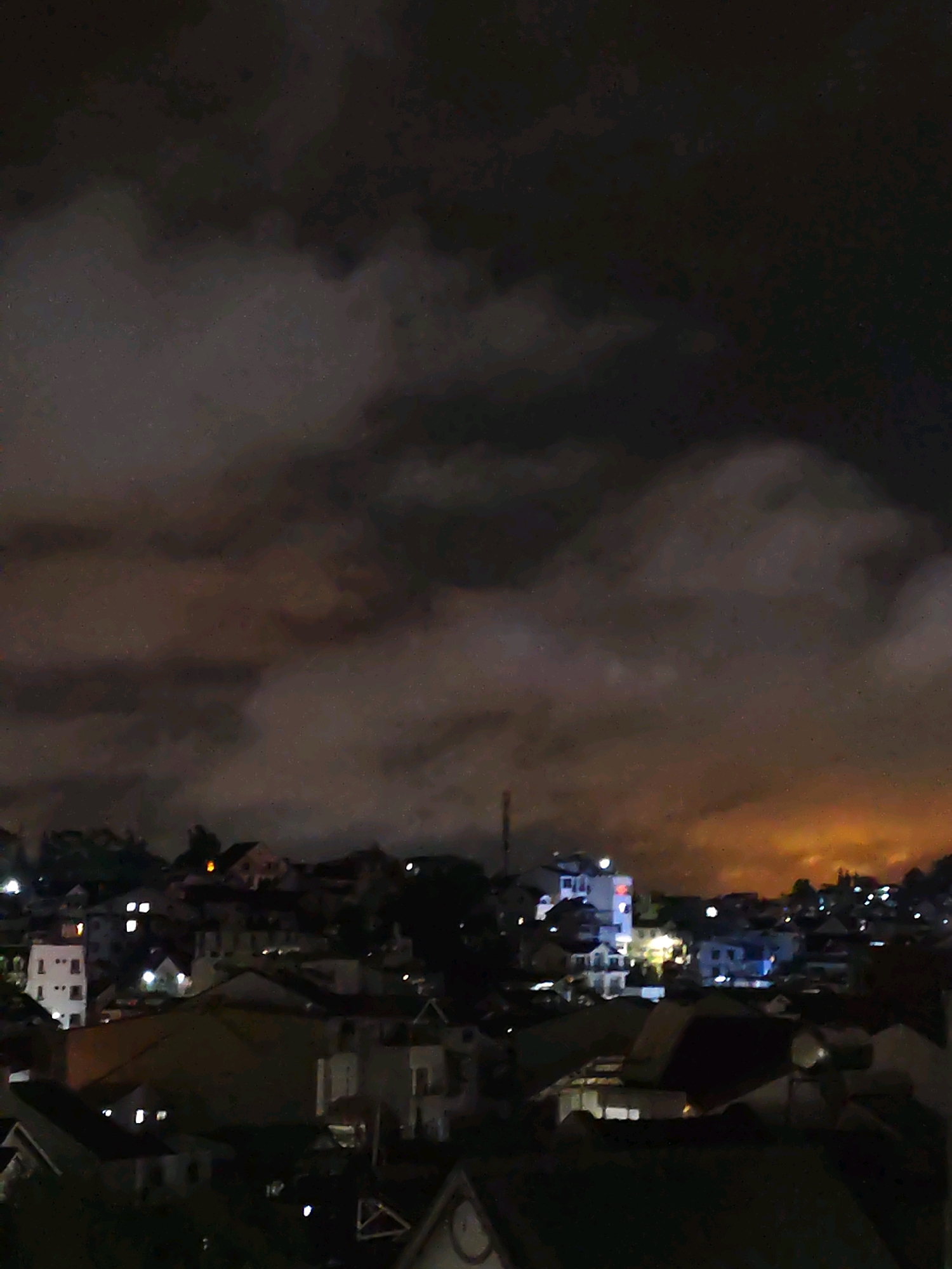 This is a nighttime aerial view of a residential area in Da Lat, Vietnam. The image is dominated by a dark, cloudy sky with diffuse, warm light emanating from the horizon, likely from distant city lights or a sunset/sunrise glow. Below the sky, a dense cluster of buildings with illuminated windows is visible, suggesting a densely populated urban or suburban landscape. The architecture appears to be primarily low-rise, with a mix of houses and apartment buildings. Some structures are illuminated with artificial lights, while others remain in shadow. The terrain seems to be hilly, with buildings built on various levels. A few streetlights or building lights are visible as distinct points of light. The overall impression is of a city at night, shrouded in clouds, with a subdued glow of artificial light.