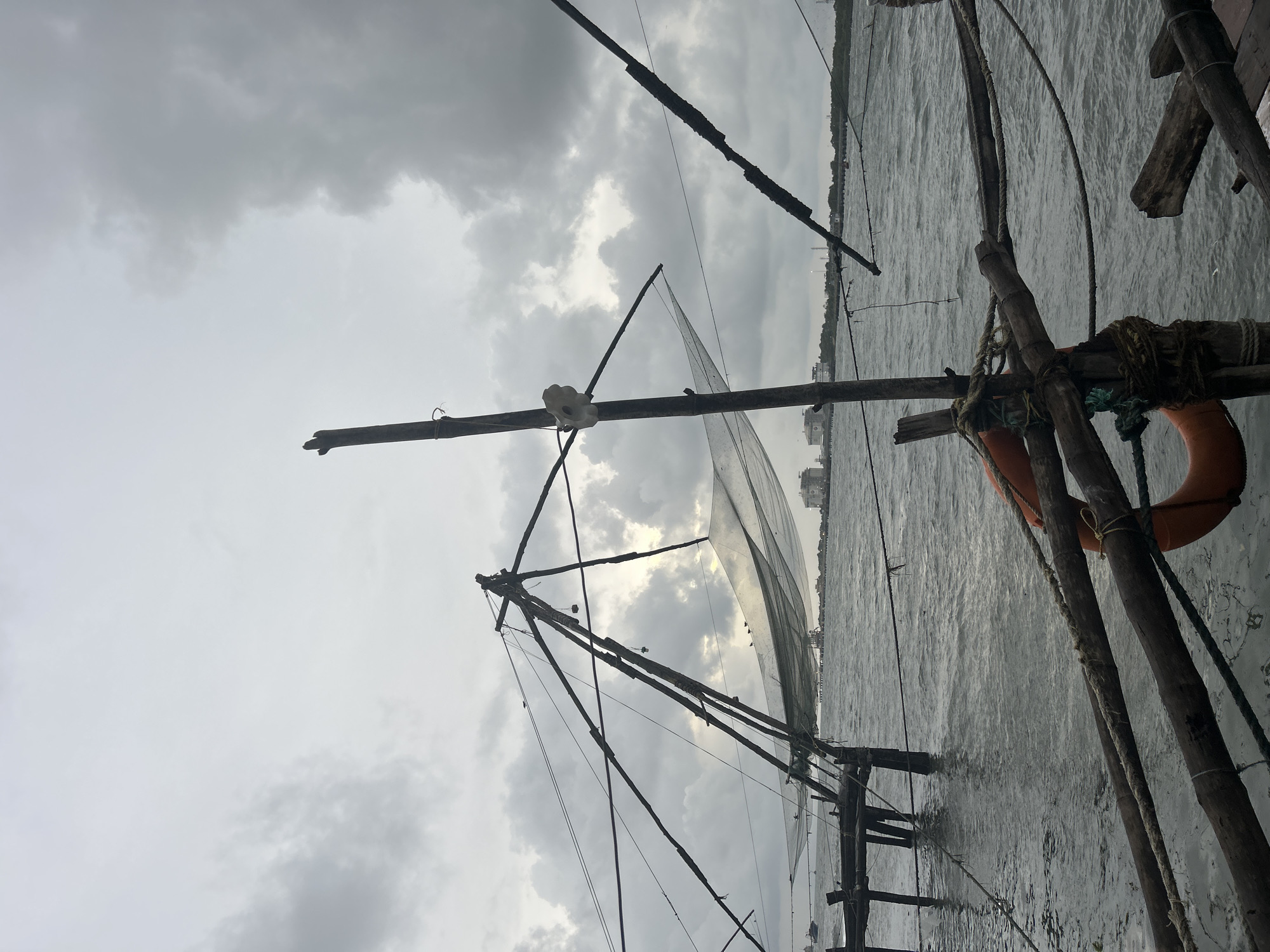 The image captures a scene featuring traditional Chinese fishing nets against a backdrop of an overcast sky and a body of water, indicative of Kochi, India. The photograph is oriented vertically (rotated 90 degrees clockwise).
The primary subjects are the large, cantilevered Chinese fishing nets, constructed from sturdy wooden poles and ropes. Two such structures are prominent, with their intricate framework silhouetted against the bright, cloudy sky. One net appears to be partially lowered into the water, while the other's complex rigging is visible. In the foreground on the right, part of another wooden structure is seen, with an orange lifebuoy tied to it, adding a touch of color to the otherwise muted palette.
The activity implied is fishing, although no specific fishing action or people are visible. The nets are set up in their characteristic manner, ready for operation.
Notable details include the weather, which is clearly overcast, with a sky full of grey clouds that provide diffused lighting. The water surface is textured with ripples, suggesting either wind or currents. In the far background, a faint outline of buildings along a distant shoreline can be observed, indicating an urban or semi-urban environment. The overall ambiance is tranquil and subdued due to the muted tones of the grey sky and water. There is no visible text in the image.