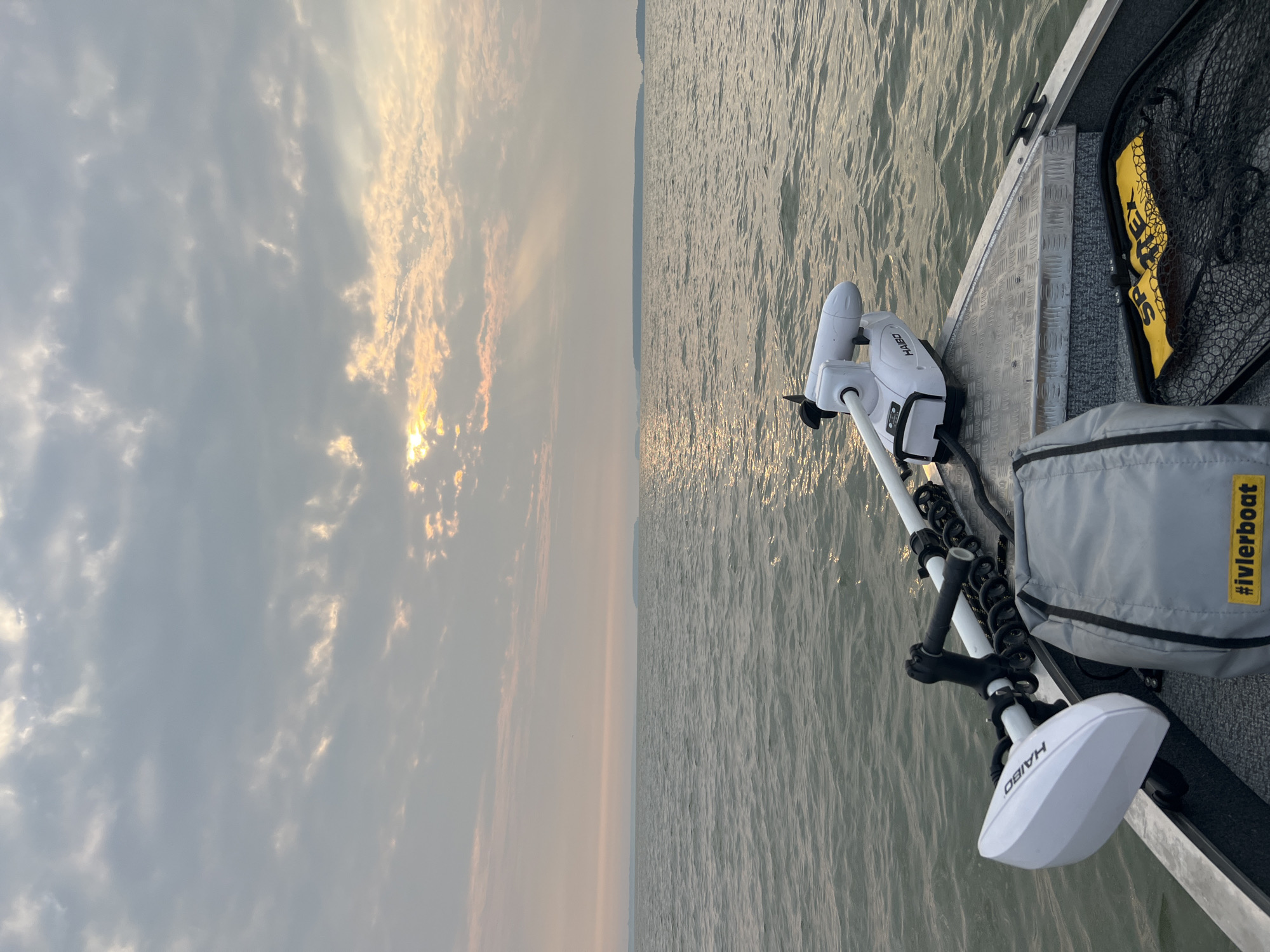 The image captures a tranquil scene from the bow of a boat on a body of water, likely during a sunrise or sunset. The sky is filled with scattered clouds, with the sun's rays piercing through, creating a gentle, diffused light. The water is calm, with subtle ripples reflecting the soft colors of the sky.
In the foreground, the boat's interior is visible, showing a trolling motor mounted on the port side. A grey bag with a yellow tag reading "#ivlerboat" is placed on the deck, next to a fishing net with two yellow objects, one of which appears to be a life vest, labeled "SPORTEX." A distant landmass, possibly an island or the shoreline, is visible on the horizon.
The image does not contain any people and no specific location in the city of Novopichugovo, Russia can be identified from the visual information provided. The scene depicts a peaceful moment on the water, suggesting a recreational activity such as fishing or leisure boating.