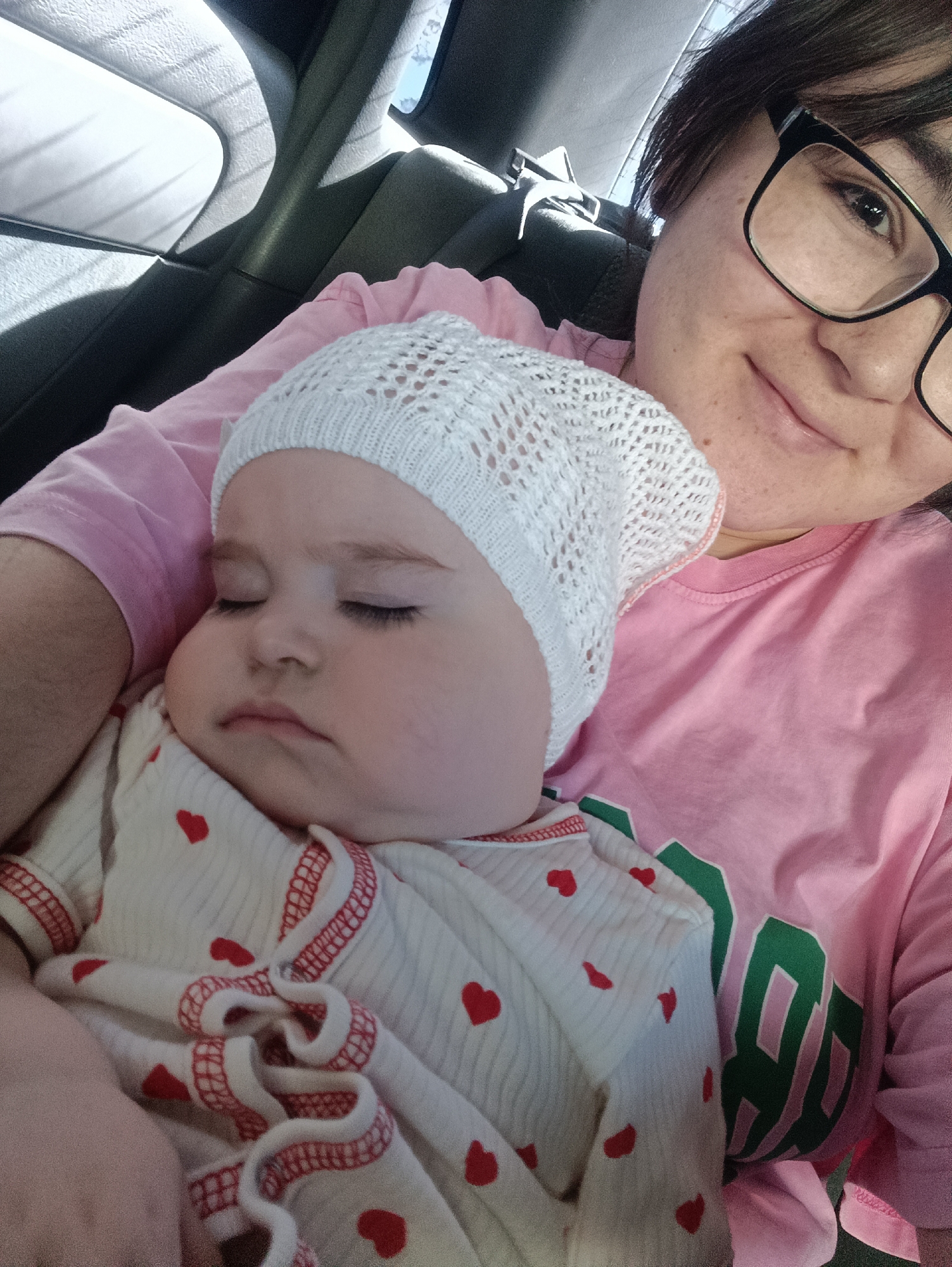 A person with black-framed glasses and dark hair is holding a sleeping baby. The baby wears a white knit hat and a white onesie with red hearts. The person holding the baby is wearing a pink t-shirt. The setting appears to be inside a car. The image was taken in the city of Chaikovsky, Russia.