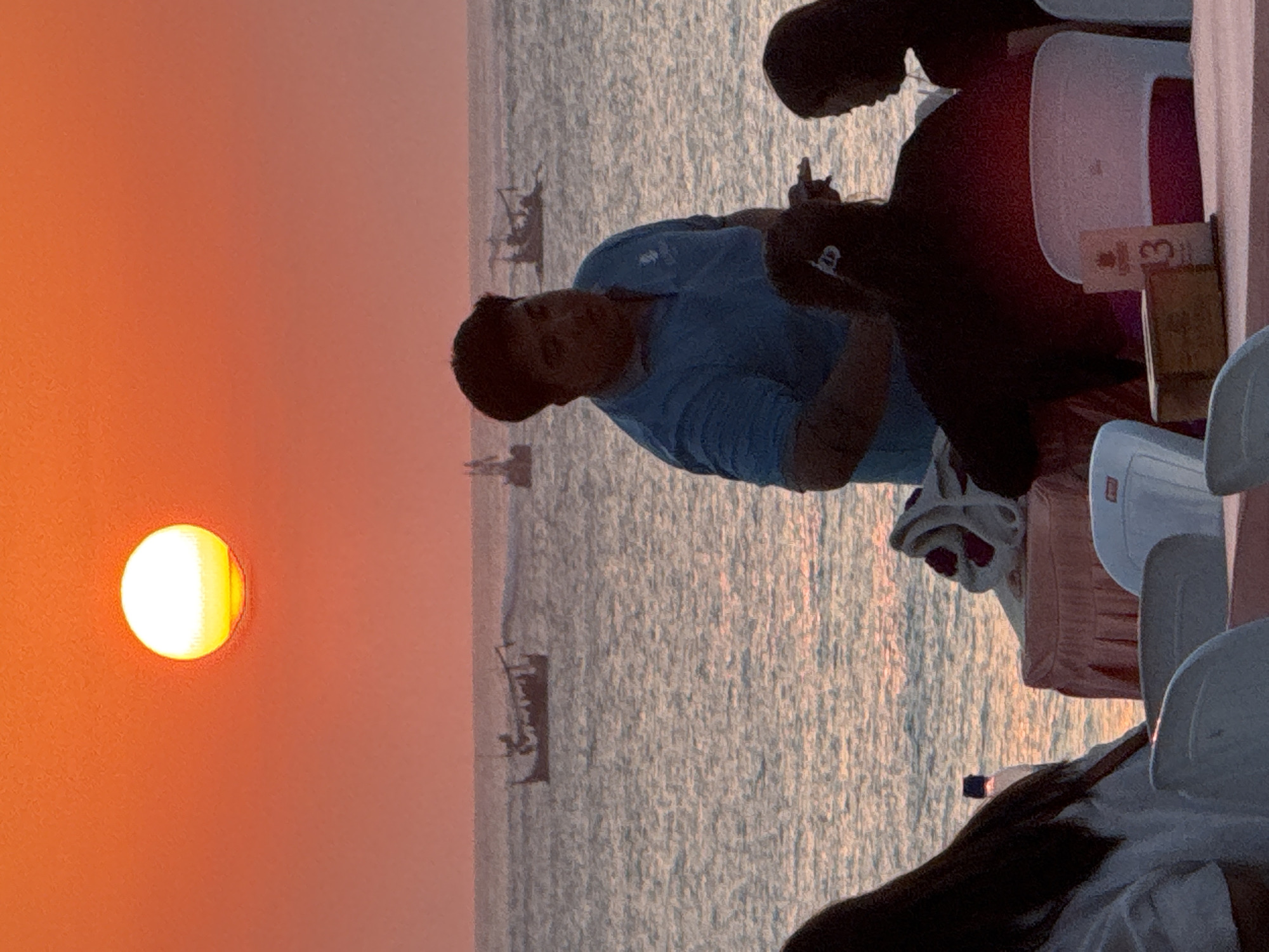An outdoor dining scene unfolds at sunset on Kuta Beach, Indonesia. The sky is ablaze with a fiery orange glow, with the sun a bright, luminous orb near the horizon. The sea stretches out, its surface rippling with the warm hues of the sunset.
In the foreground, three people are partially visible. To the left, a person with long dark hair faces away from the camera, wearing a white top. In the center, a man in a blue polo shirt stands, looking towards the right. He appears to be interacting with a person seated to his right, whose back is to the camera, wearing dark clothing and with dark hair. A third person, a woman with dark hair, is visible to the far right, facing left.
A table is set with what appears to be a pink tablecloth. On the table, a small wooden box and a table number displaying "3" are visible. White plastic chairs surround the table. In the middle ground, several boats are present on the water, silhouetted against the sunset. Two of these appear to be traditional fishing vessels. The overall atmosphere suggests a relaxed dining experience by the sea.