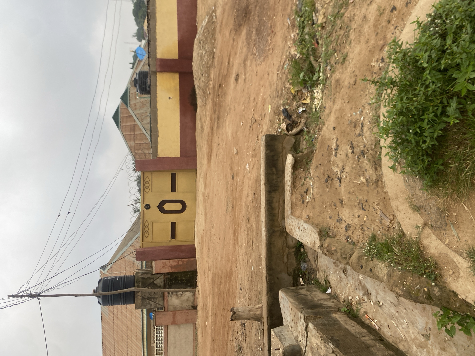A horizontal, medium shot shows the exterior of a property in Barden Yana, Nigeria. The scene is overcast and appears to be a residential area. In the foreground, a concrete ditch with some light vegetation and debris runs from the bottom left corner diagonally towards the center of the image. Behind the ditch is a dirt path with a small pile of light brown dirt on the right side of the frame. A weathered, grey wooden stump sits in the dirt path. Dominating the midground is a yellow metal gate with a dark brown outline of the number "0" in the center, flanked by two dark brown vertical lines. The gate is set within a wall constructed of yellow and maroon sections, with barbed wire running along the top. To the left of the gate, a tall, black water tank is visible atop a concrete structure, adjacent to a corrugated metal roof with a lighter brown hue. Behind the wall, more corrugated metal roofs of varying shades of brown and green are visible. A light pole with numerous electrical wires extends from the left side of the frame towards the top center. The overall environment appears somewhat unkempt, with visible debris in the ditch and on the ground.