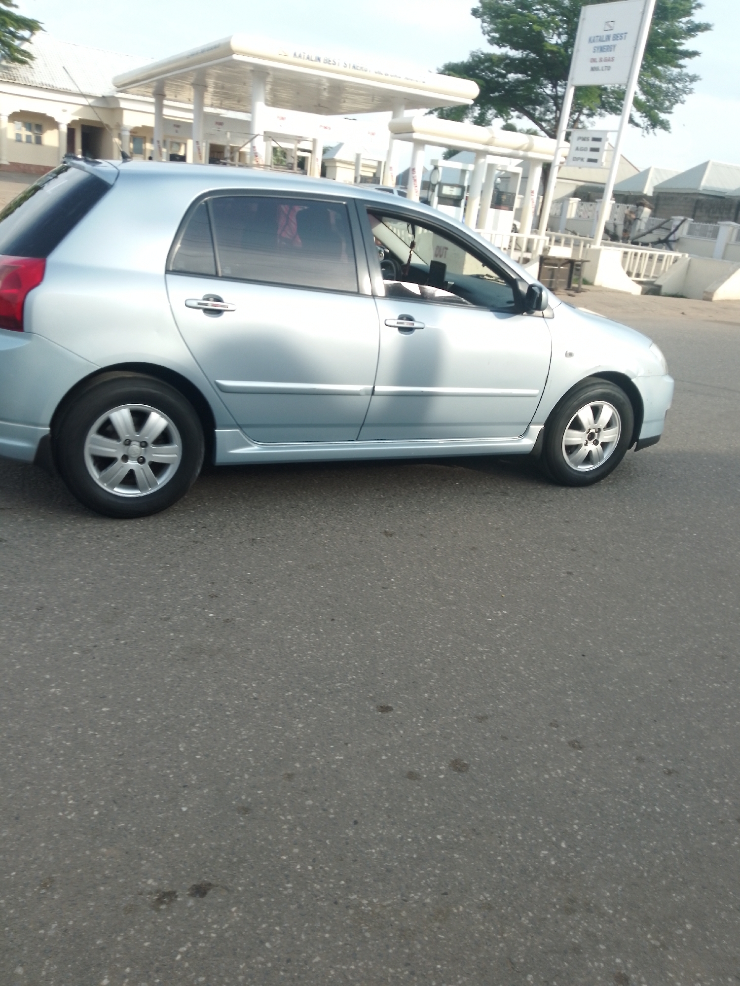 In Serti, Nigeria, a silver hatchback car is parked on an asphalt road in front of a gas station. The gas station has a white canopy supported by pillars, and a tall sign displaying "KATALIN BEST SYNERGY OIL & GAS NIG. LTD." The sign also lists fuel prices, including "PMS," "AGO," and "DPK." The car is positioned on the right side of the frame, facing left. The road surface is dark gray with visible white speckles. The background shows some buildings and trees. The sky above is bright and appears to be daytime.