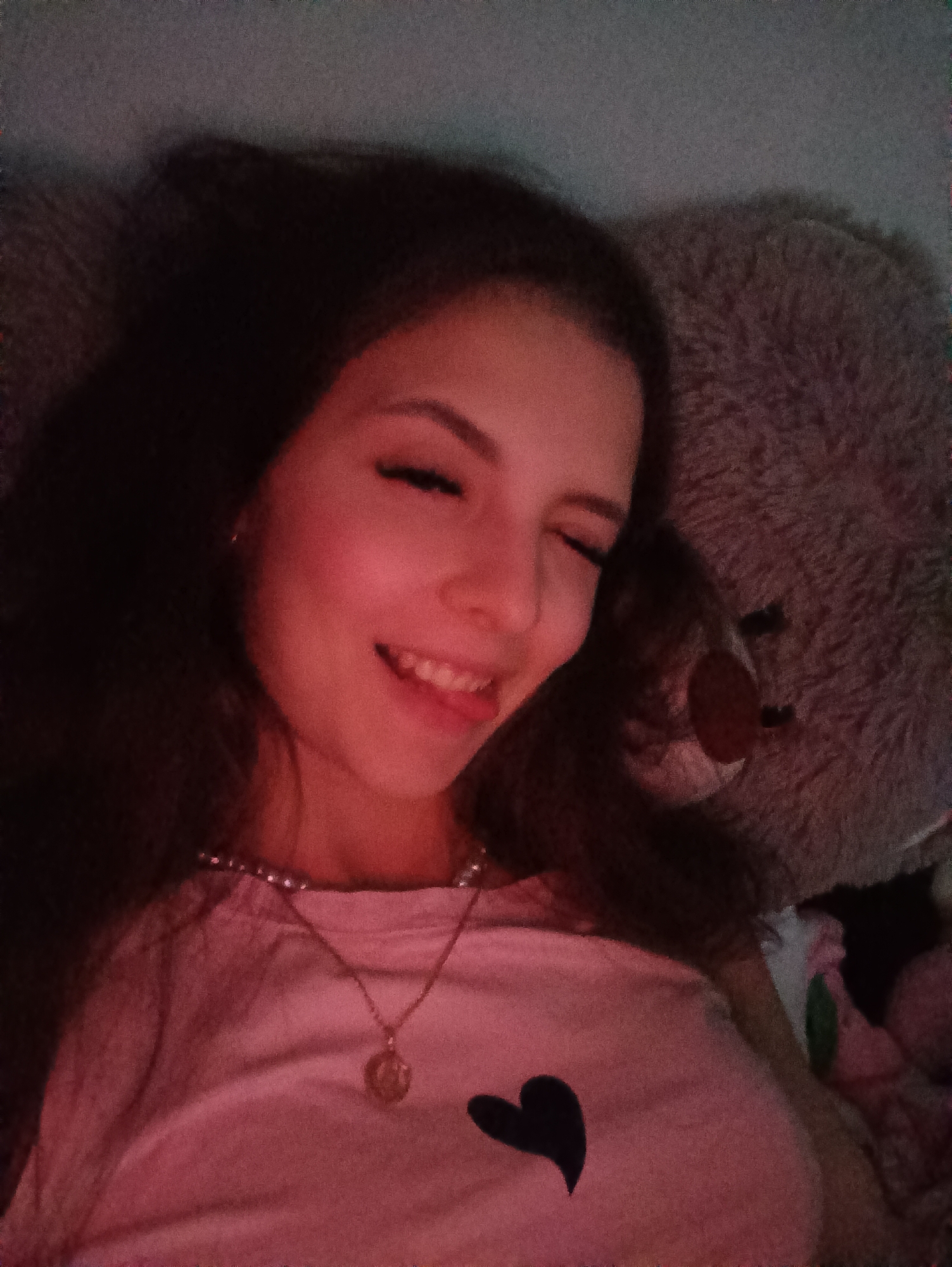 A person is lying on a pillow or cushion with a large, stuffed bear beside them. The person is smiling and appears to be wearing a pink shirt with a black heart. The subject has long, dark hair and is wearing a necklace. The location is indoors, likely in a private residence. There is no indication of what city or other external location that this image has been taken.