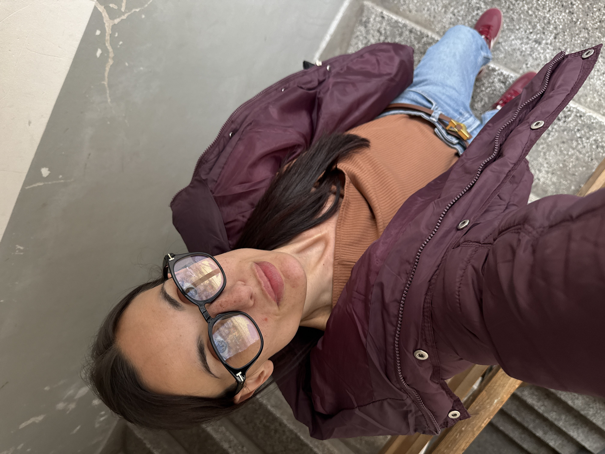 A woman takes a selfie on a staircase. She wears black framed glasses, a brown turtleneck, light blue jeans with a brown belt and gold buckle, a maroon puffer jacket, and maroon shoes. The staircase is concrete with gray steps. The location could be Pleven, Bulgaria but there is no clear indication of the city visible in the image.