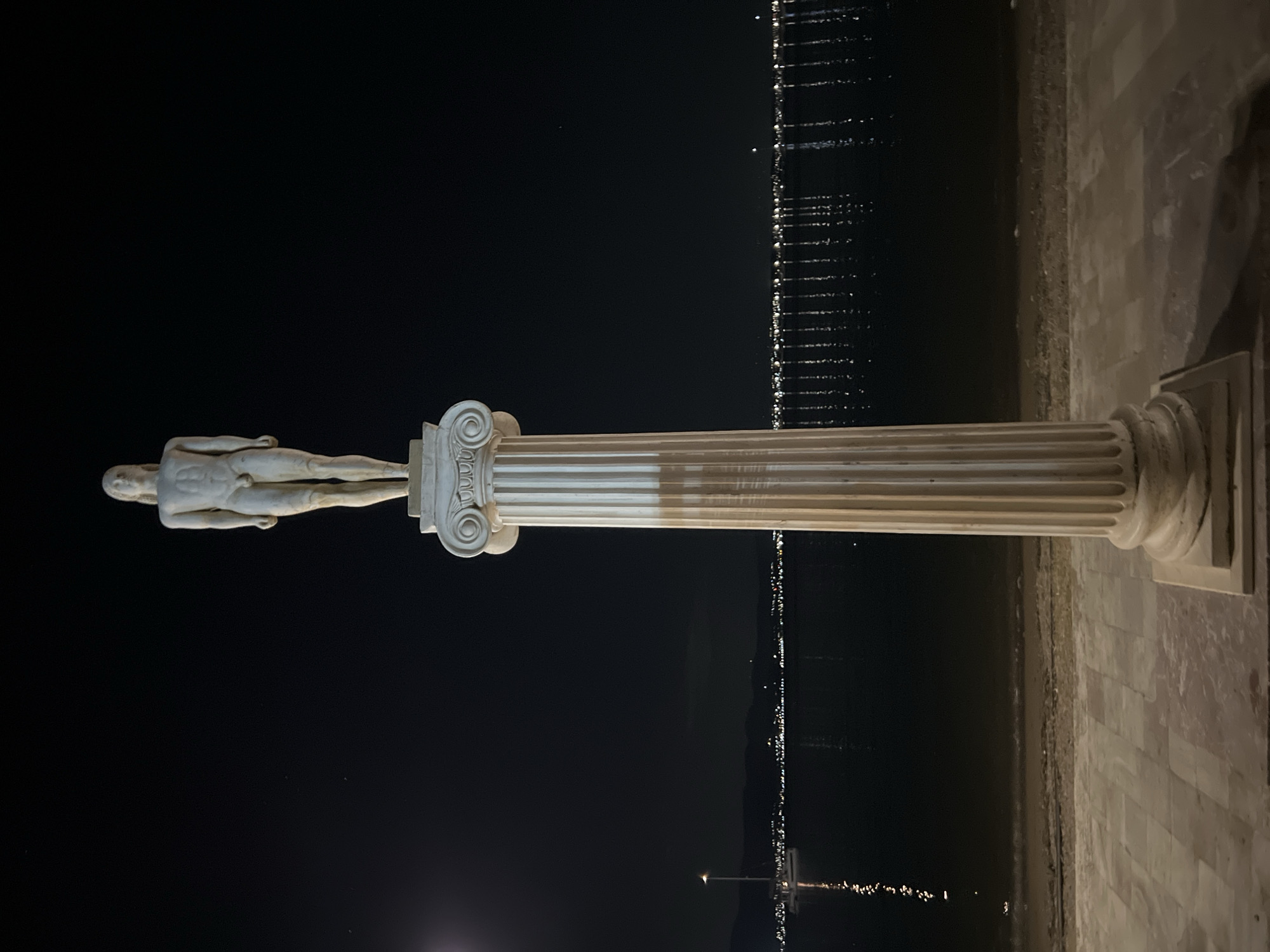 A statue of a male figure stands atop a fluted column at night in Eretria, Greece. The statue, carved from a pale stone, depicts a nude male with long hair, a muscular physique, and a circumcised penis. The column itself is also pale stone, with prominent vertical fluting and an ornate, scrolling capital. The statue and column are positioned on a paved walkway. In the background, a dark body of water, likely the Aegean Sea, stretches out, reflecting the lights of a distant city or town that lines the shore. A single sailboat with a light post is visible on the water to the left. The sky is dark and clear, with a few faint stars visible. The overall scene is serene and illuminated by artificial light sources.