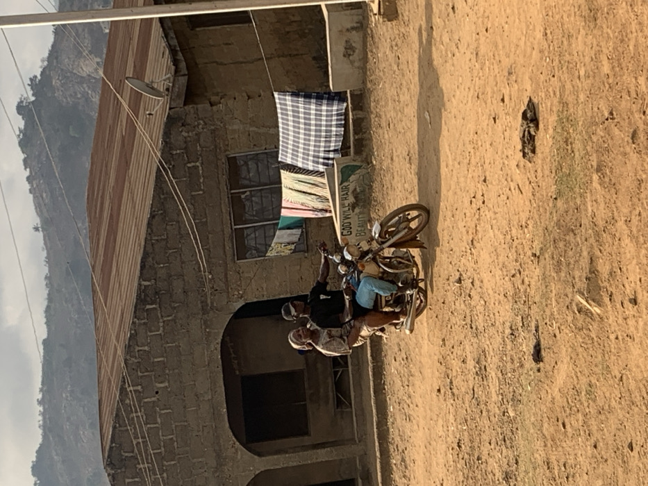 Three individuals ride a motorcycle on a dirt road. A building with a rusty metal roof, a satellite dish, and clothes hanging outside stands to the left. A sign for "God Will Hair Beauty" is also visible. The background features a mountainous landscape partially obscured by a hazy sky. The scene appears to be set in a rural or less developed area. The image is rotated to the side.