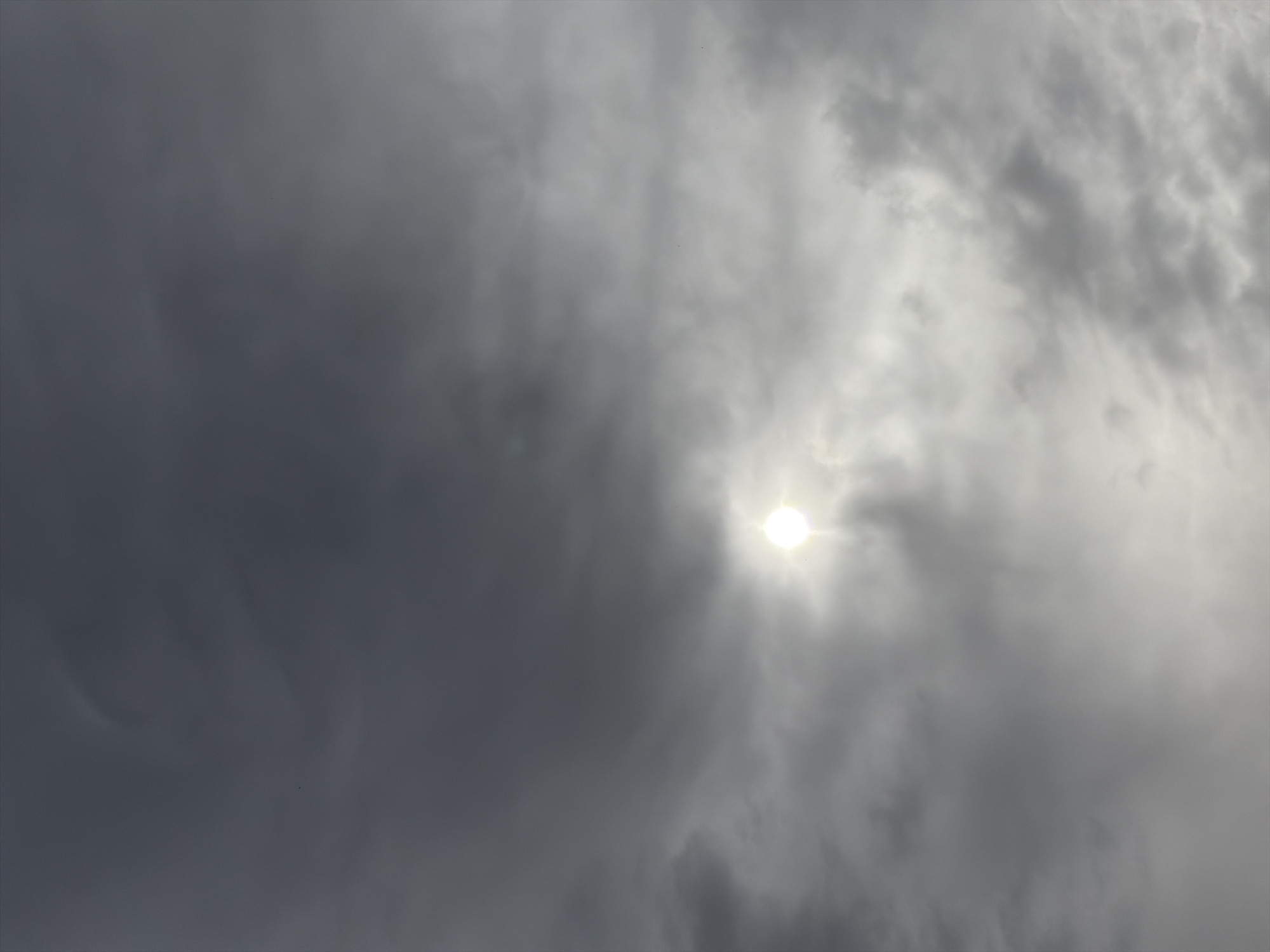 The image depicts a cloudy sky with the sun shining weakly through the overcast. The clouds are varying shades of gray, with the darkest concentrations on the left side of the image. The sun appears as a bright, indistinct orb in the center-right portion of the sky. The location is unknown.