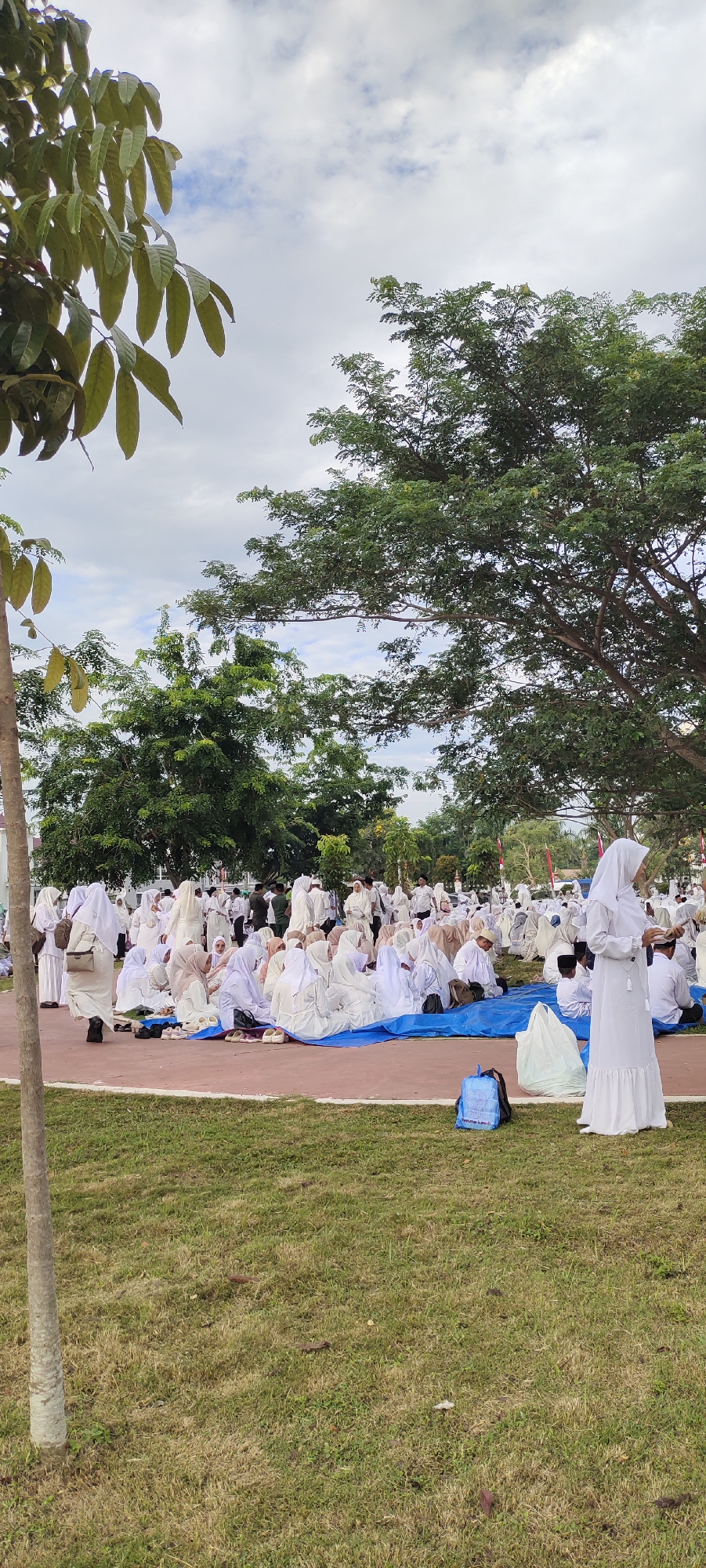 In Meunasahaluedrienlhoksukon Barat, Indonesia, a large group of people, primarily women dressed in white Islamic attire including hijabs, are gathered outdoors on a grassy area with some blue tarps spread on the ground. They are seated, facing in various directions, and appear to be participating in a religious or community event. Some individuals are standing, including a woman in the foreground on the right wearing a white dress and hijab, holding a small book. In the background, more people are visible, along with trees and a cloudy sky. The environment suggests an open park or public space.