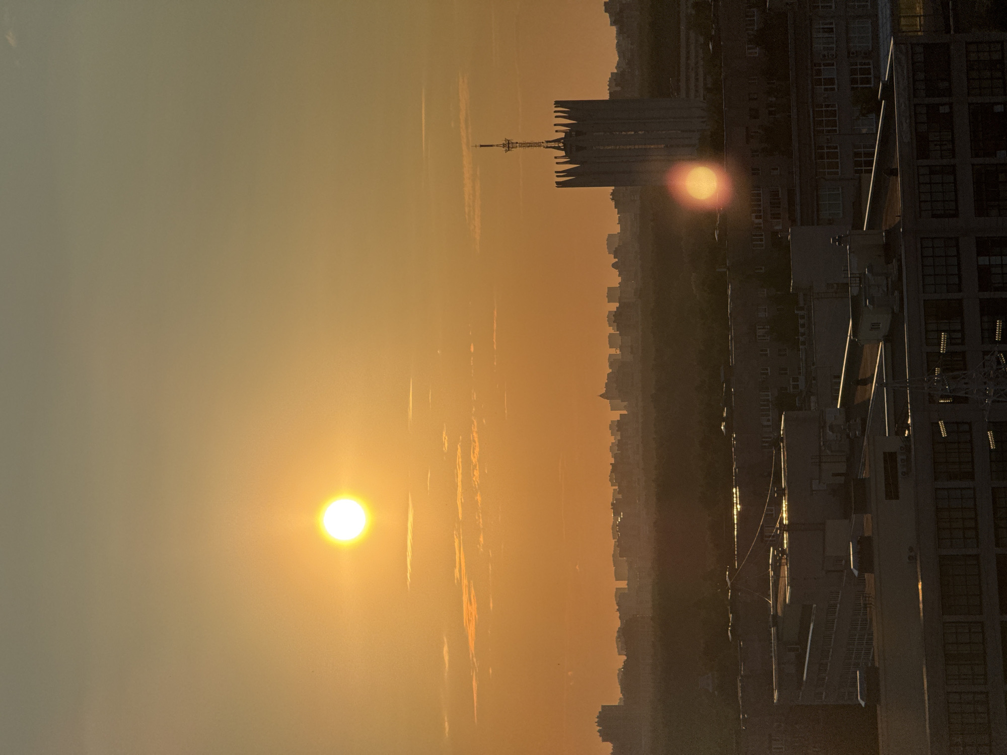 The image depicts a sunset over a city skyline in Unknown, Russia. The sun, a bright orb, is positioned in the upper left quadrant of the image, casting a warm orange glow across the sky and the urban landscape below. Wispy clouds are visible, tinged with the hues of the setting sun.
Dominating the middle ground is a distinctive, tall, white building with a pointed spire topped by a cross. This structure is flanked by rows of lower-rise buildings, characteristic of a city. In the foreground, industrial or commercial buildings with flat roofs and numerous windows are visible, appearing somewhat silhouetted against the brighter sky. The overall impression is of a sprawling urban environment bathed in the atmospheric light of sunset. There are no people or animals visible in the scene.