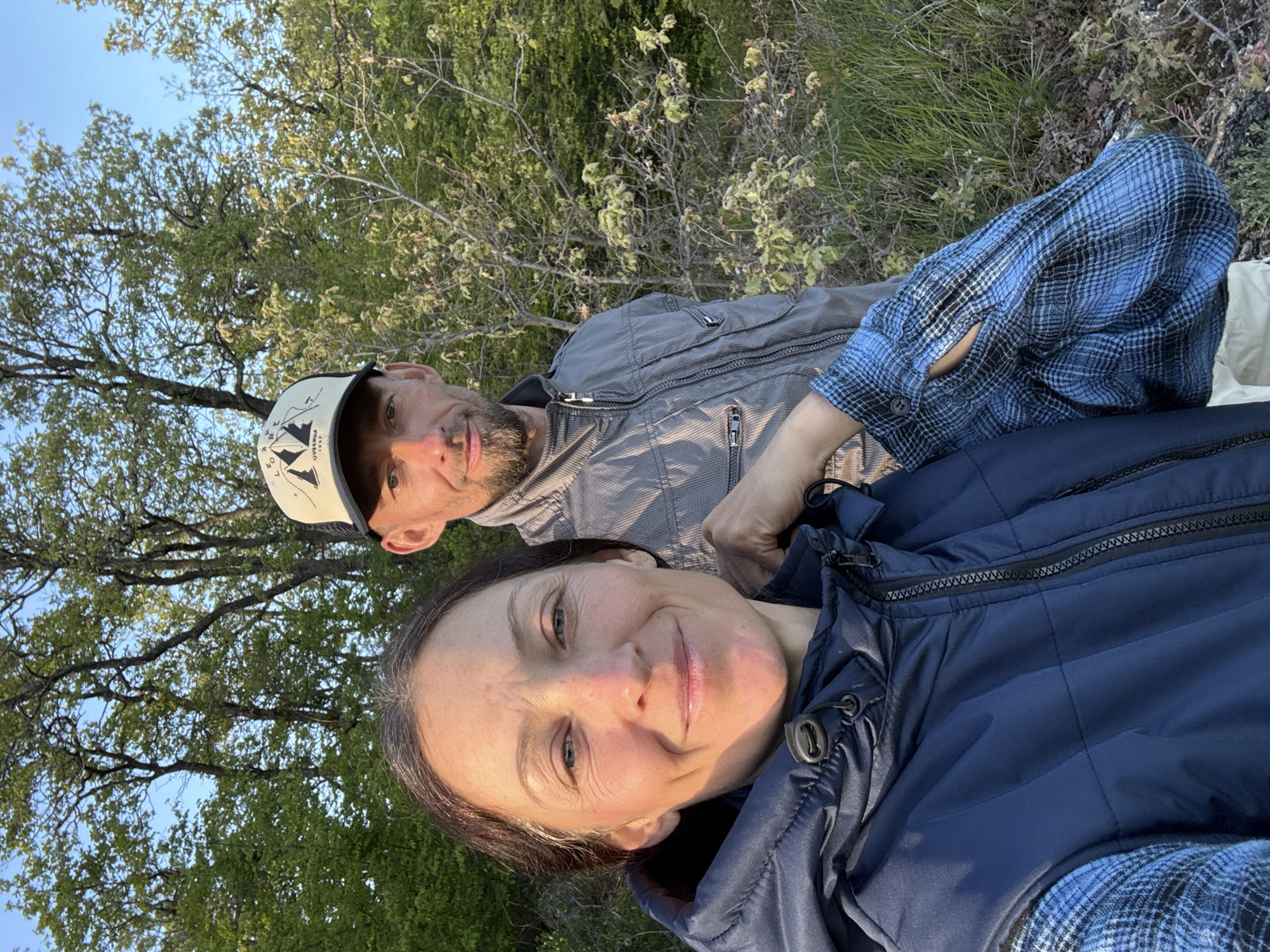A low-angle, outdoor photograph depicts two individuals in Kiparisnoe, Ukraine. The woman in the foreground wears a navy blue quilted jacket and smiles at the camera. Behind her to the left is a man with a light beard wearing a beige baseball cap with an image of mountains on it and a gray zip-up jacket. They appear to be in a wooded area with trees and foliage visible in the background. The sky is partly visible through the trees and is a light blue color.