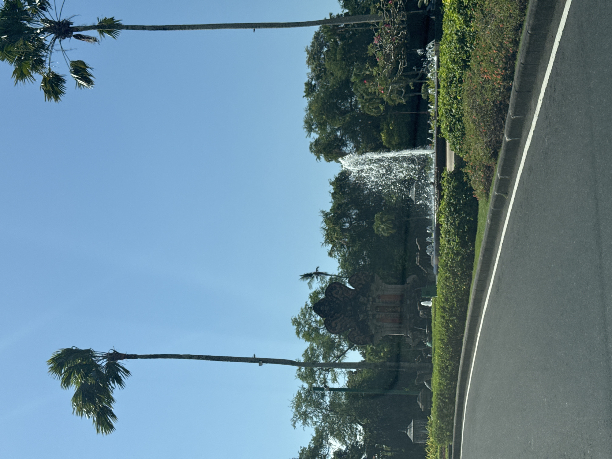 In Kuta Selatan, Indonesia, a road curves in the foreground, bordered by a manicured green lawn and a curb with a white line. Beyond the lawn, a fountain sprays water upwards, with a decorative Balinese-style structure behind it. Two tall palm trees stand on either side of the fountain, with lush green trees forming the backdrop under a clear blue sky. The scene is captured from a moving vehicle.