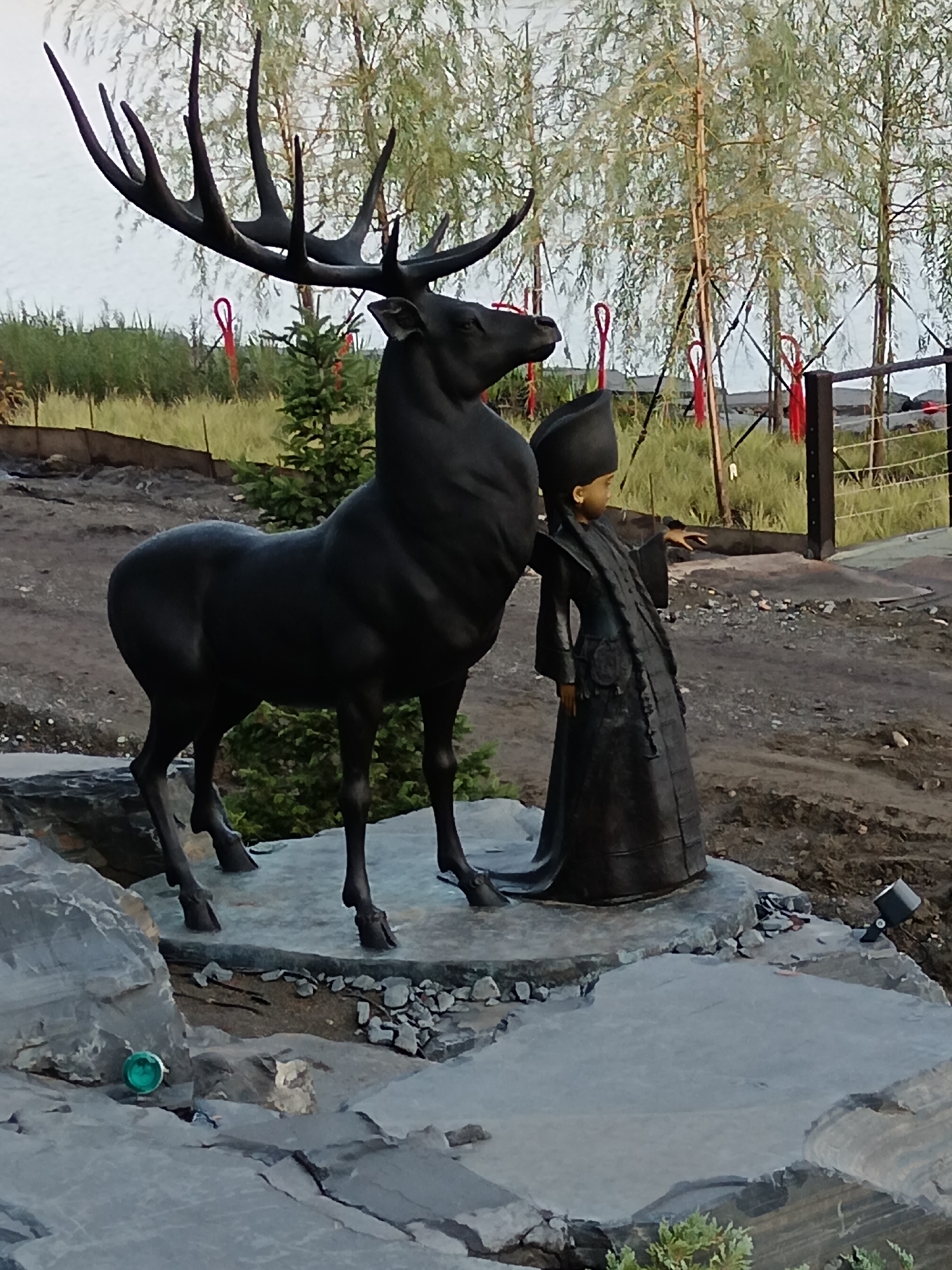 The image depicts a bronze sculpture of a reindeer and a child located in Ozernoe, Russia. The sculpture is positioned on a rocky outcrop with trees in the background. The reindeer is dark bronze, with large antlers and a muscular build. It stands to the left of the child. The child is also bronze, wearing traditional clothing including a tall hat, a long robe, and a sash. The child's right hand is extended towards the reindeer. In the foreground, there are large, grey rocks. To the left, on one of the rocks, a green, circular object resembling a glass ball or a decorative sphere is visible. The background shows a fence with red decorations, vegetation, and a body of water in the distance. The lighting suggests it is daytime.