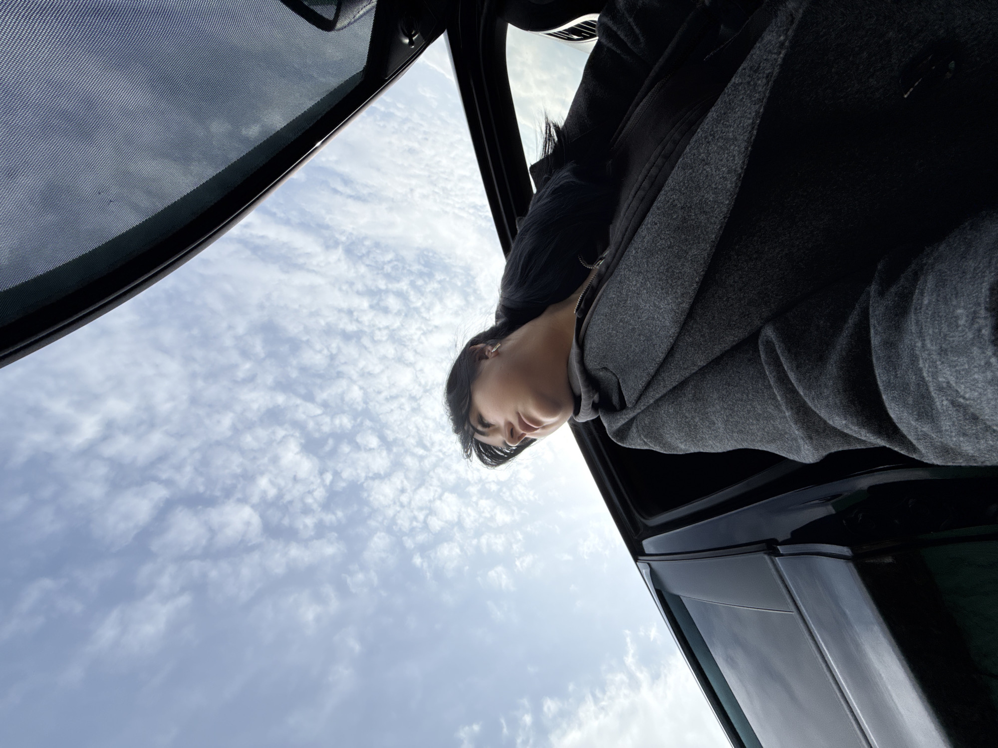 A low-angle shot shows a woman looking down from a car. She has dark hair and is wearing a gray coat. The sky is visible through the open car window, with wispy clouds scattered across the blue. The interior of the car is black. There are no visible landmarks to indicate a specific location in Chaikovsky, Russia. The scene appears to be focused on the woman's perspective and her interaction with the open window and the sky.